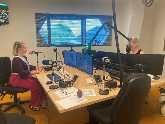 Two women in a radio broadcast studio with microphones, computer monitors, and recording equipment. One woman is sitting at the table, and the other is seated opposite her. Large windows show a cloudy sky and trees outside.