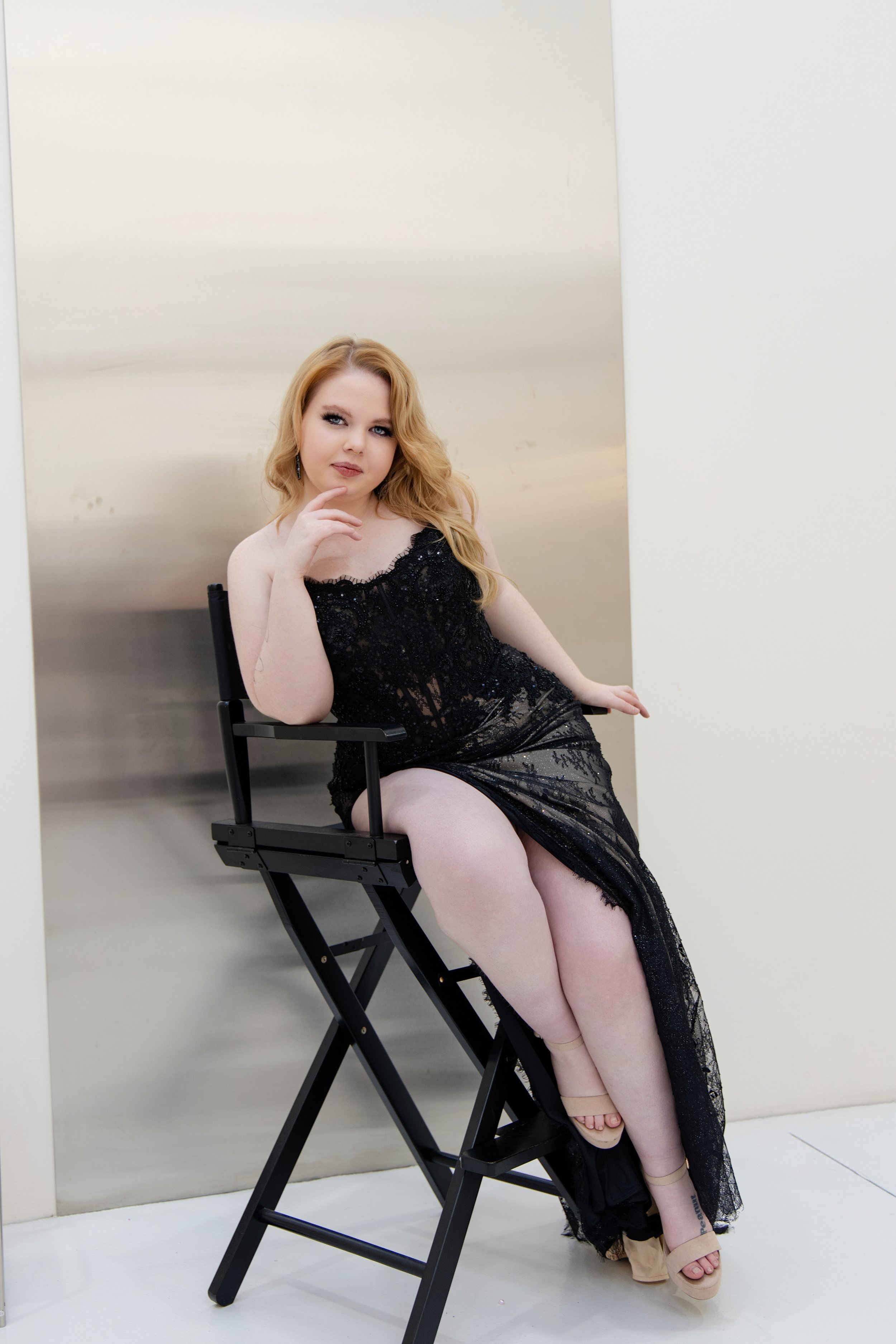 A woman with red hair sitting on a director's chair, wearing a black lace dress with a slit, beige shoes, and posing with her hand near her face in front of a minimalist background.