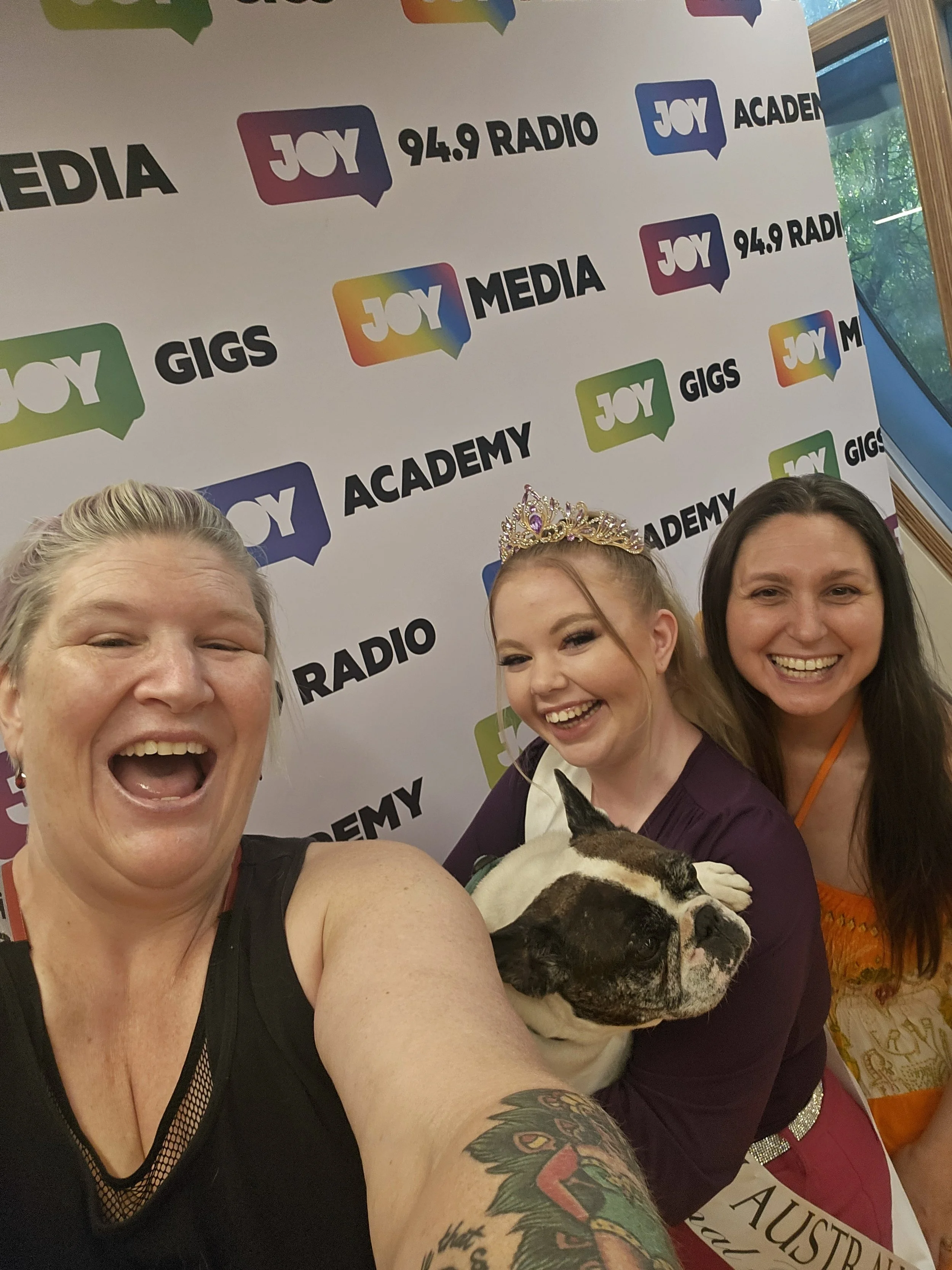 Three women smiling for a selfie with a Boston Bulldog puppy, against a backdrop with logos of Joy Media, Gigs, and Joy 94.9 Radio.