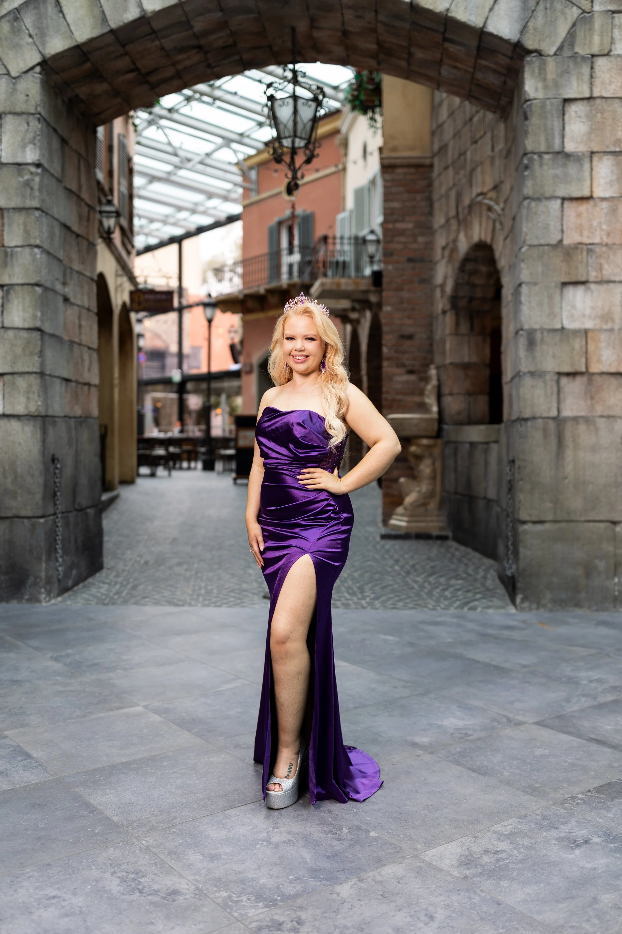 A blonde woman wearing a purple strapless gown with a high slit, a tiara, and high heels, standing outdoors in front of a stone archway and brick building, smiling at the camera.