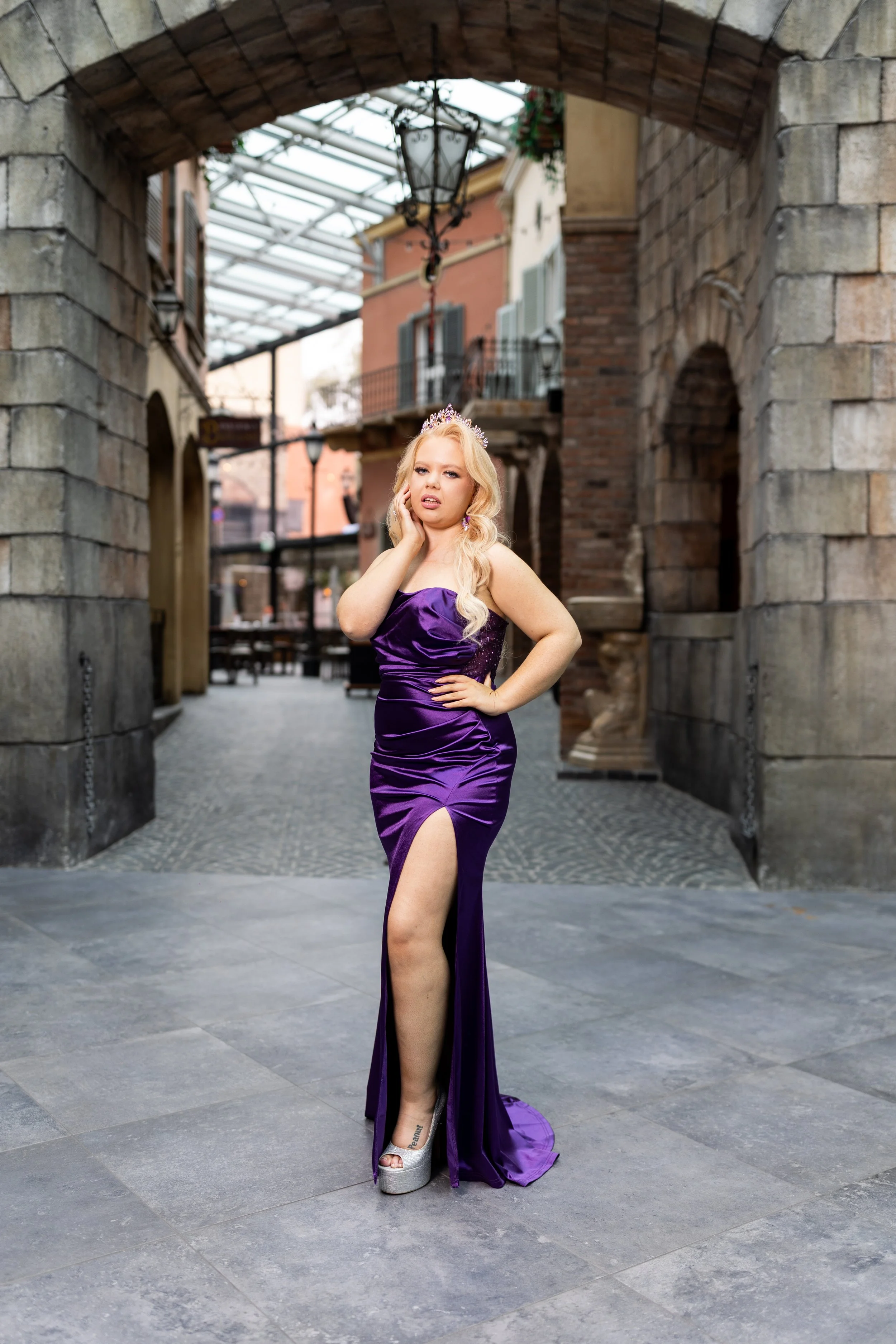 A woman with long blonde hair wearing a purple satin dress with a high slit, a tiara, and silver platform heels, standing under an arched stone gate in an urban setting with buildings and balconies in the background.