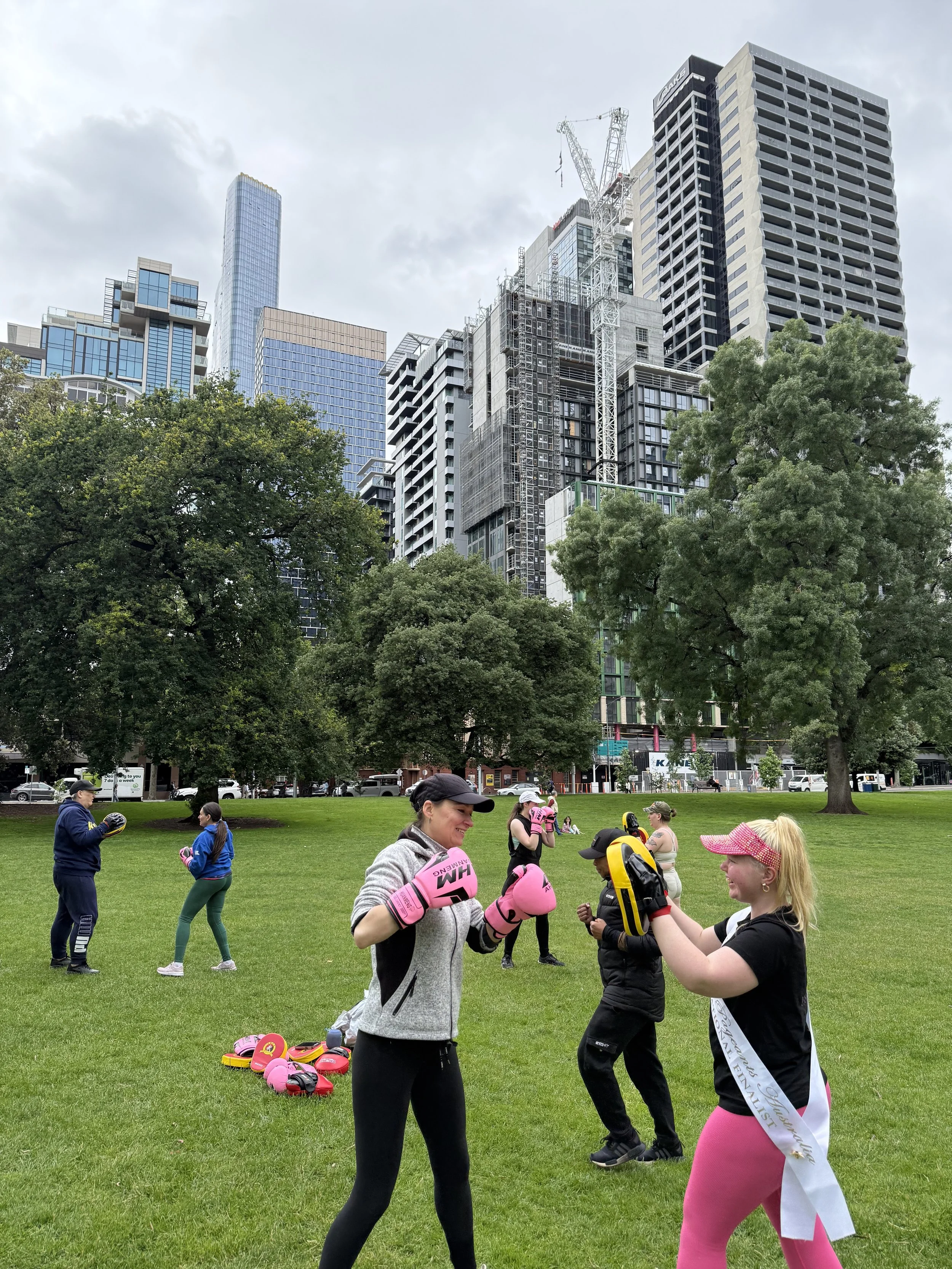 A group of women and girls are practicing boxing outdoors in a park with green grass and trees, with tall modern buildings in the background.
