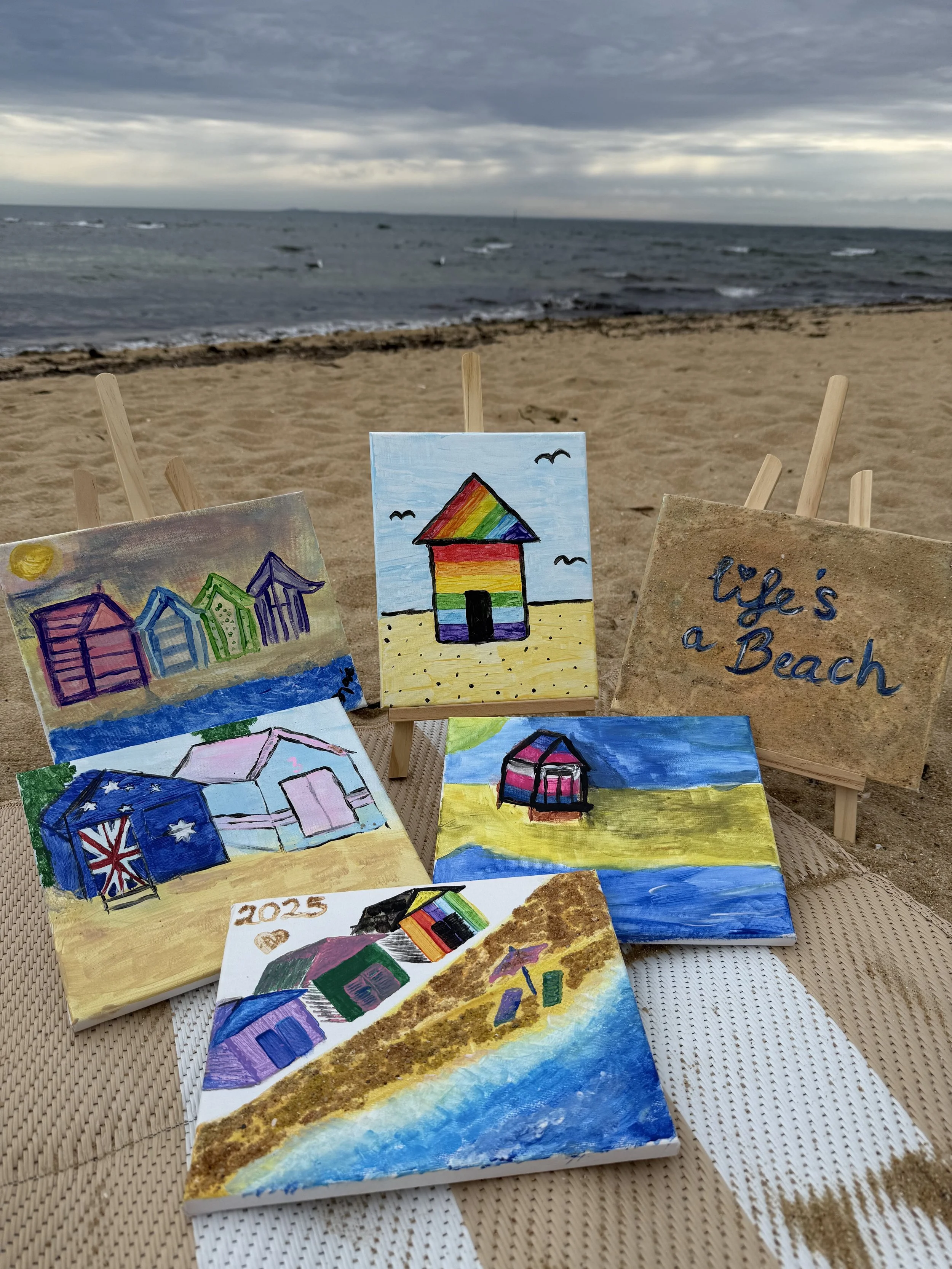 A collection of colorful beach-themed paintings displayed on small wooden easels on the sand with the ocean and cloudy sky in the background.