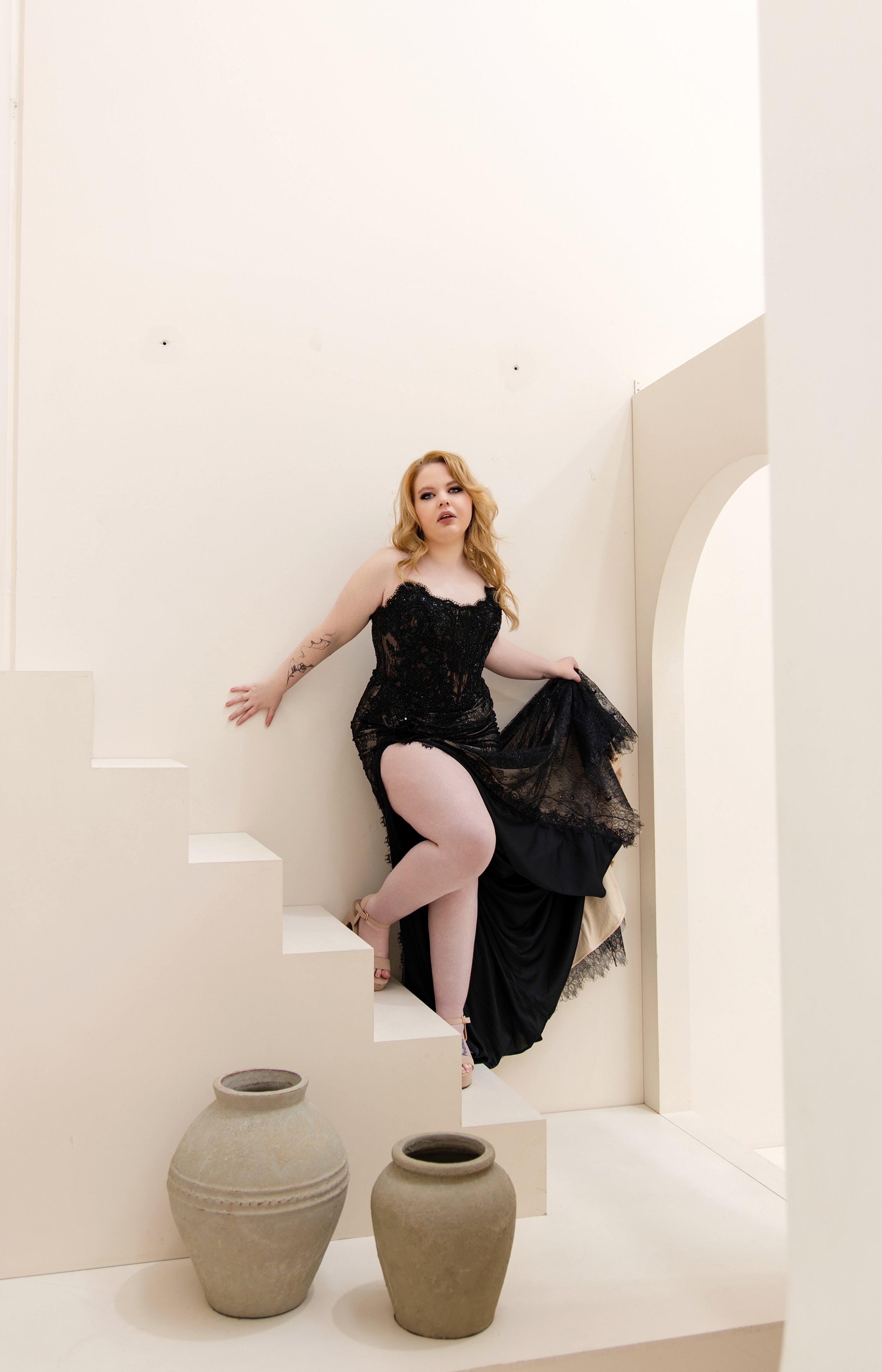 A woman with red hair wearing a black lace dress, posing on a staircase with her hand on the wall and one leg raised, against a plain white wall with large, beige pottery vases on the floor.