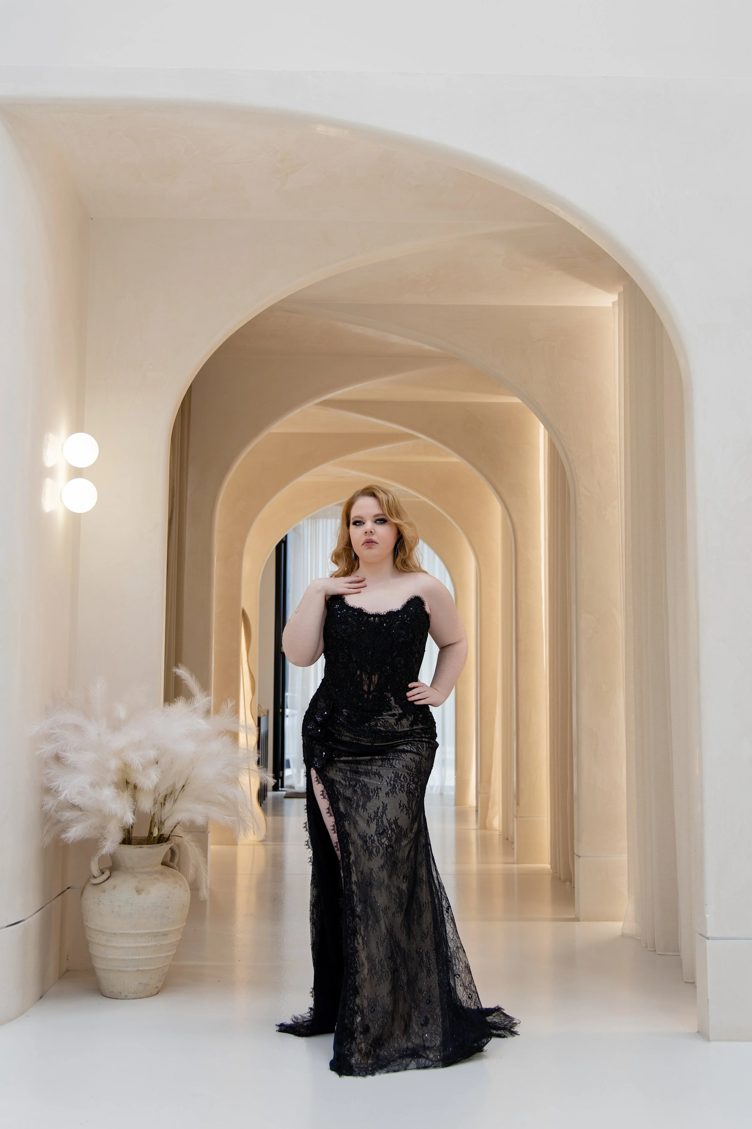 A woman in a long, black lace wedding gown standing in a modern, cream-colored hallway with curved archways.