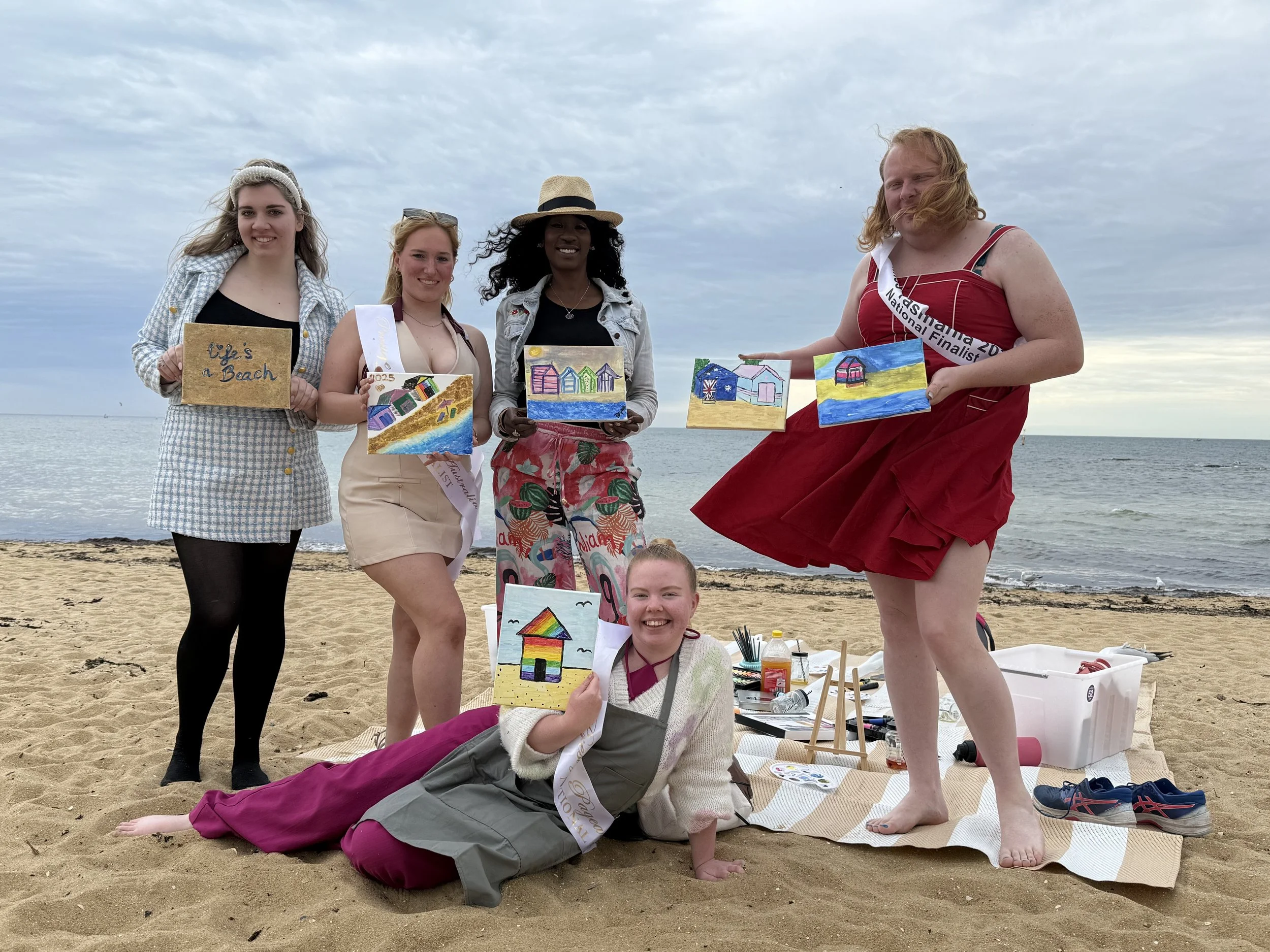 Group of five women and a girl at the beach, some holding or displaying handmade art projects of colorful beach huts, with art supplies and shoes on the sand, cloudy sky in the background.