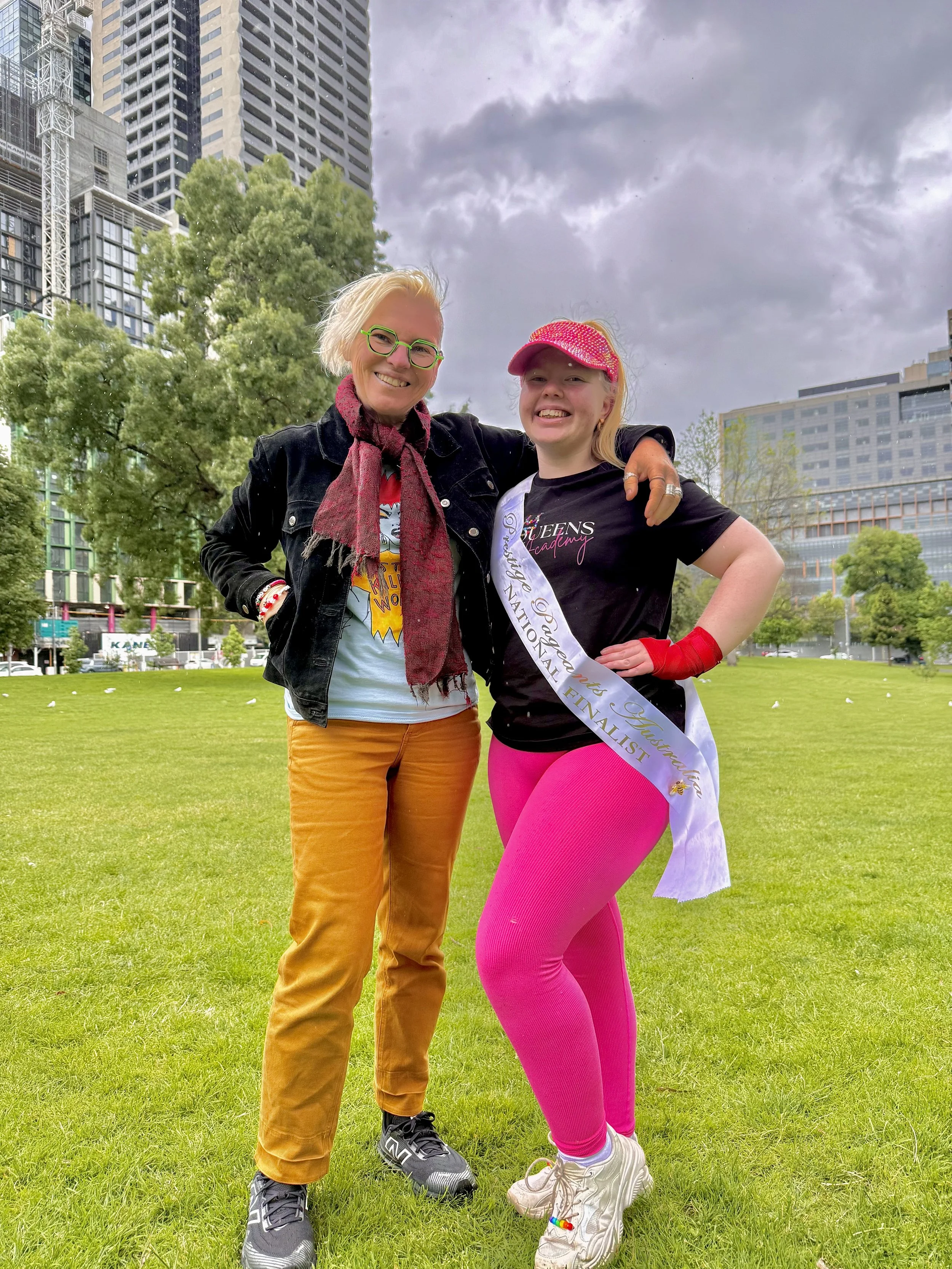Two smiling women standing on grass in an urban park, with tall buildings and trees in the background. One woman has short blonde hair with green glasses, wearing a black jacket, a scarf, and mustard-colored pants. The other woman has blonde hair wit