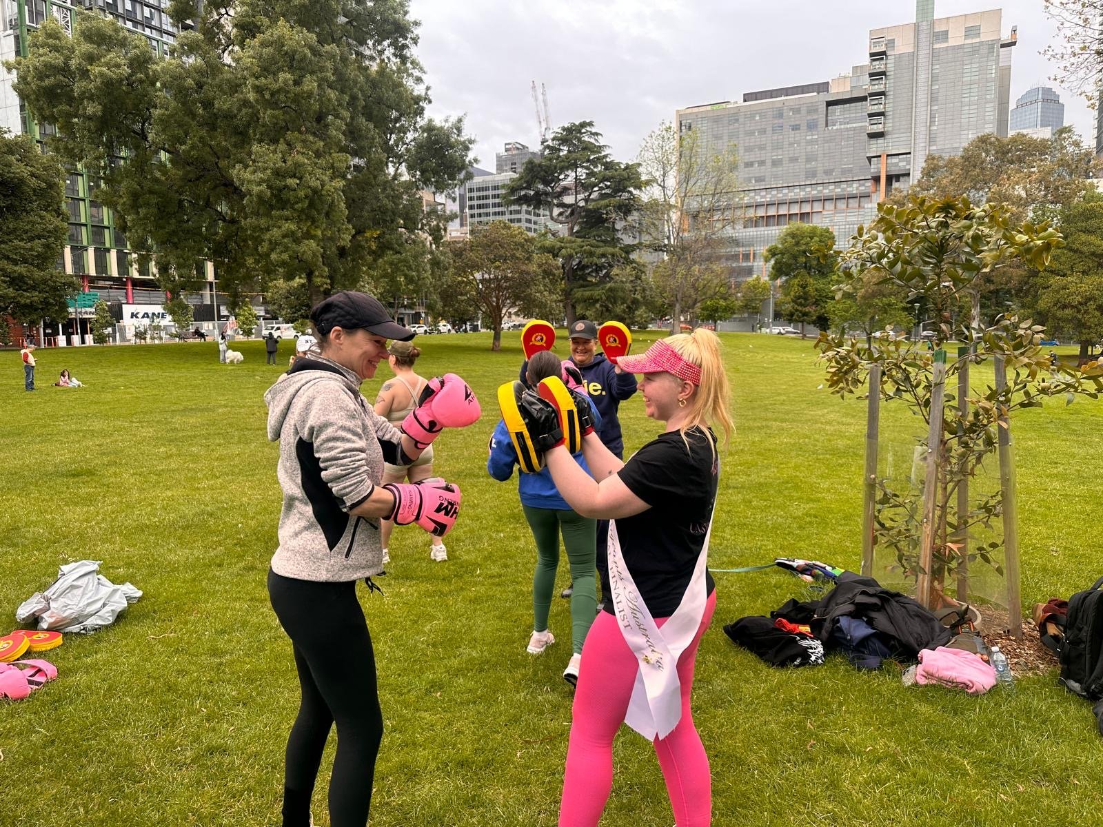 Women practicing boxing outdoors in a park with a group of people, trees, and city buildings in the background.