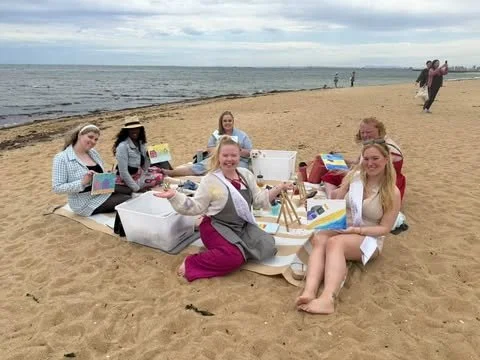 Group of women and girls having a picnic on the beach with bags and food, near the ocean under a cloudy sky.
