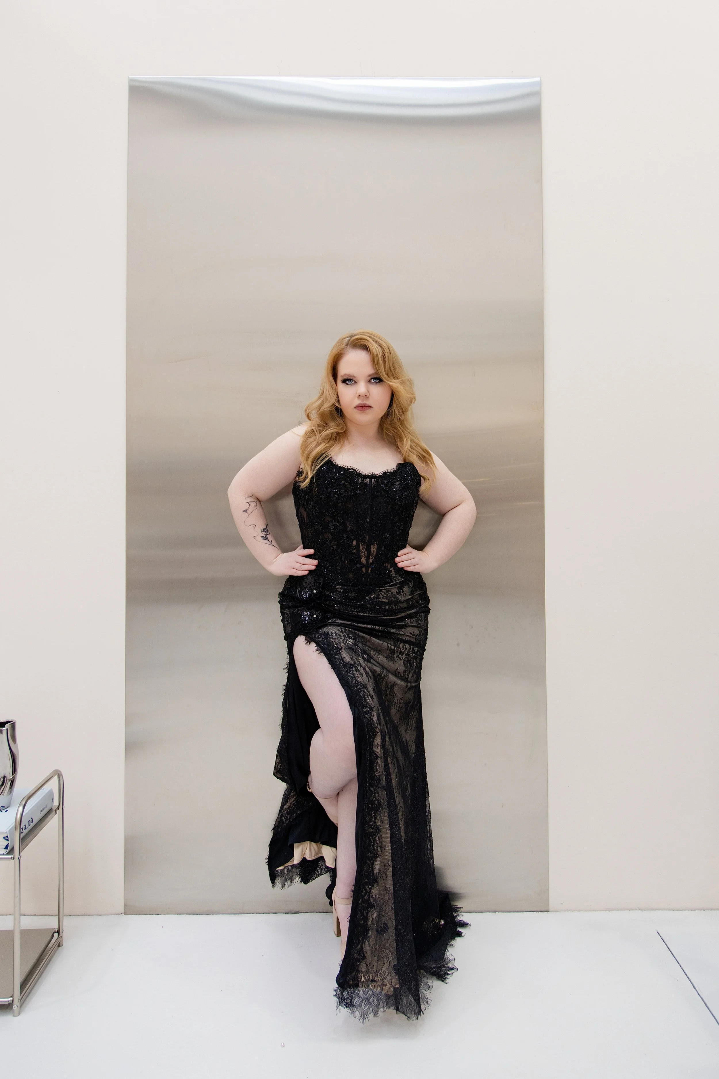 A woman with wavy blonde hair and light makeup stands with her hands on her hips in front of a metallic silver backdrop, wearing a black lace dress with a thigh-high slit and beige tights.