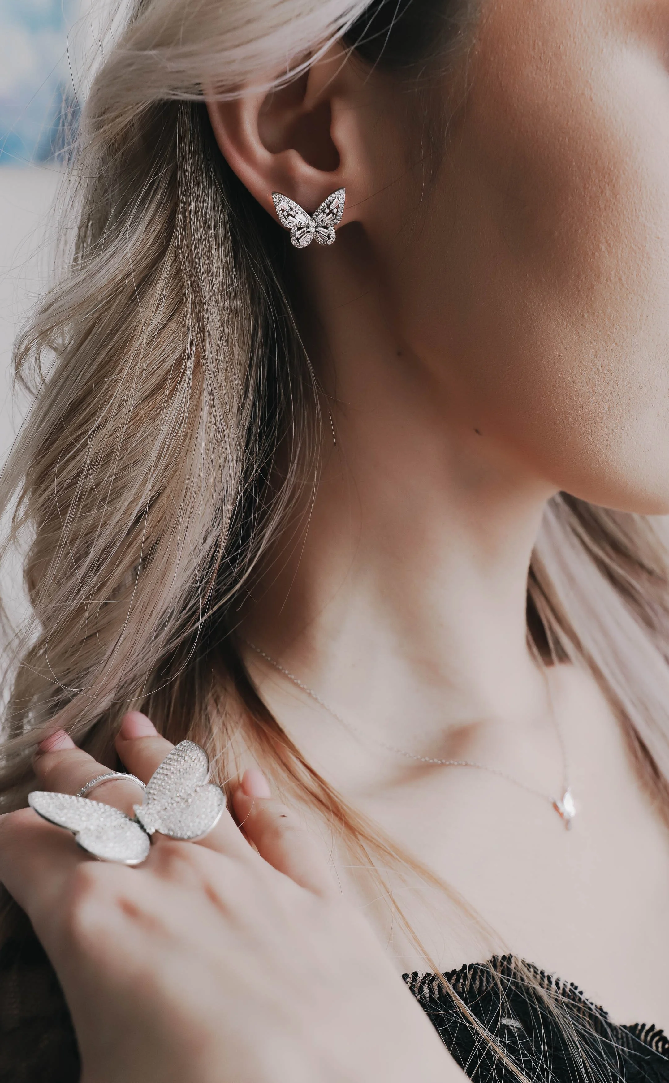 Close-up of a woman's ear with butterfly-shaped diamond earrings, jewelry on her fingers, and a delicate necklace. She has wavy blonde hair and light skin.