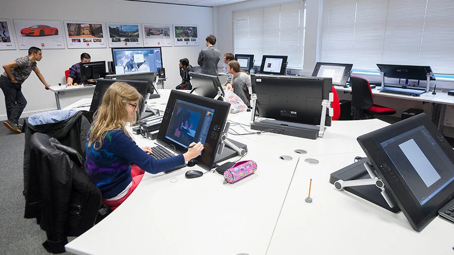 A classroom or studio with students working on digital art or animation projects on drawing tablets and computer monitors. Some students are seated, while others stand or walk around. The room has large windows and framed pictures on the wall.