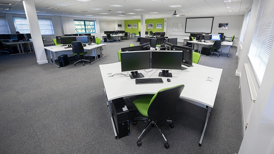 Empty modern office space with multiple desks, each equipped with dual monitors, keyboards, and office chairs. The room has windows with blinds, green accent walls, and a large white presentation screen at the front.