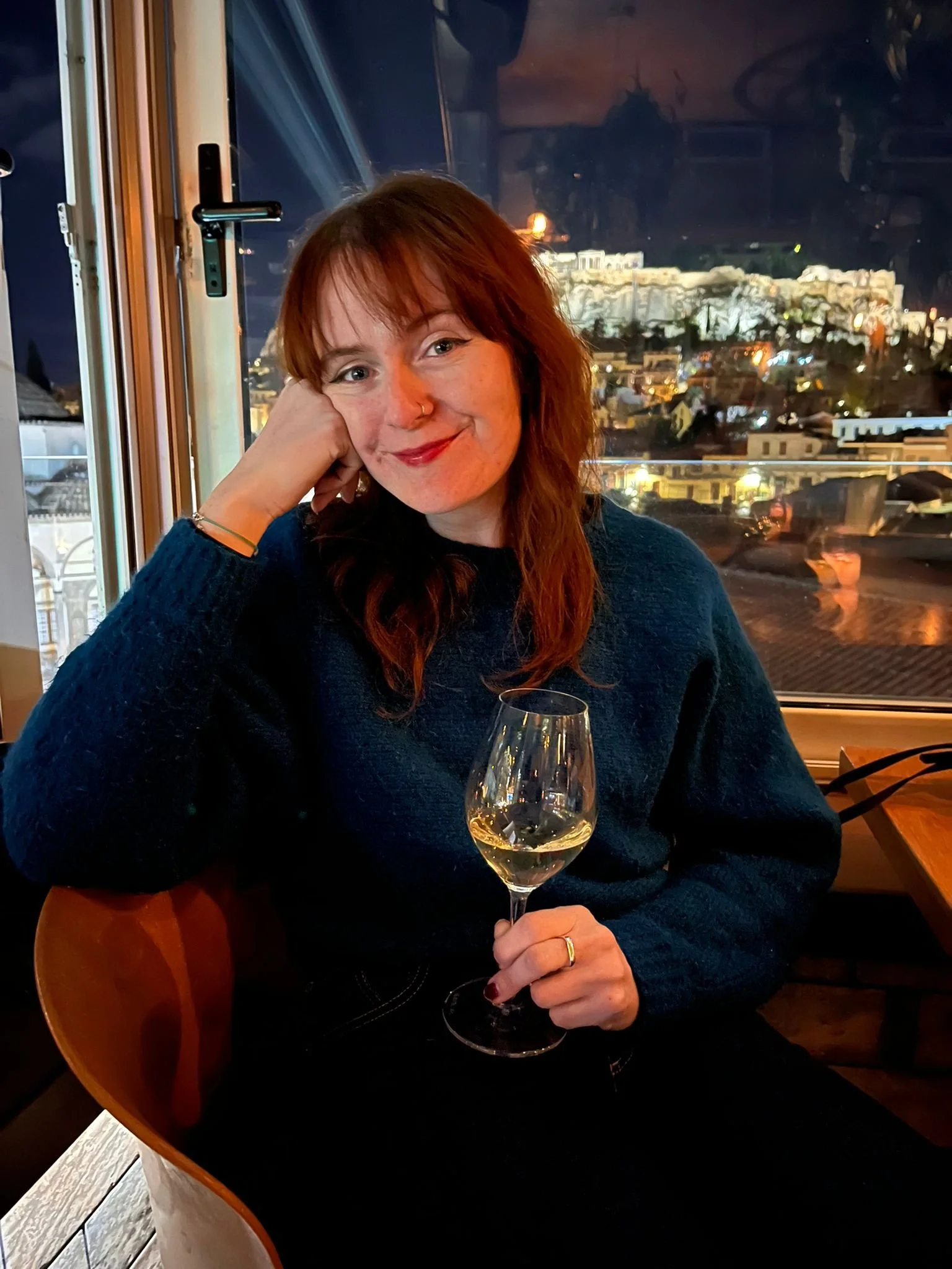 A woman with red hair and a nose ring sitting at a table with a glass of white wine, smiling, in a restaurant near a window with a night cityscape view.