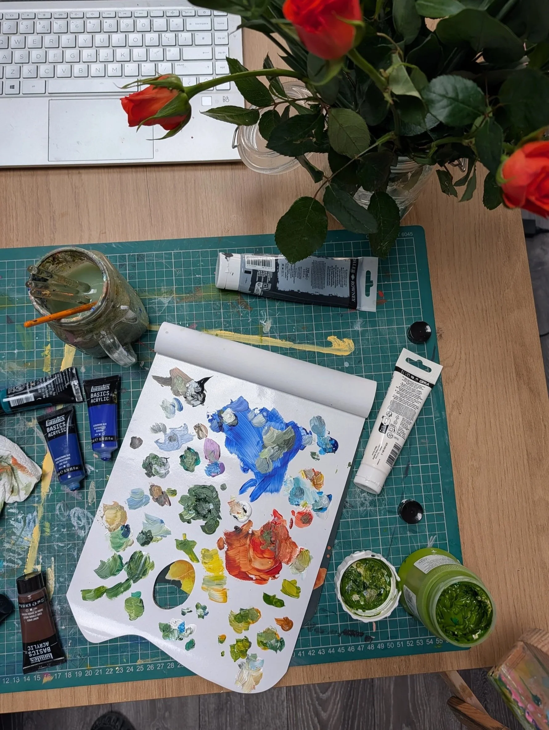 A table with art supplies including a palette with various paint colors, paint tubes, a jar with brushes, a laptop, a potted plant with red flowers, and a work surface with paint smudges and a painted paper.