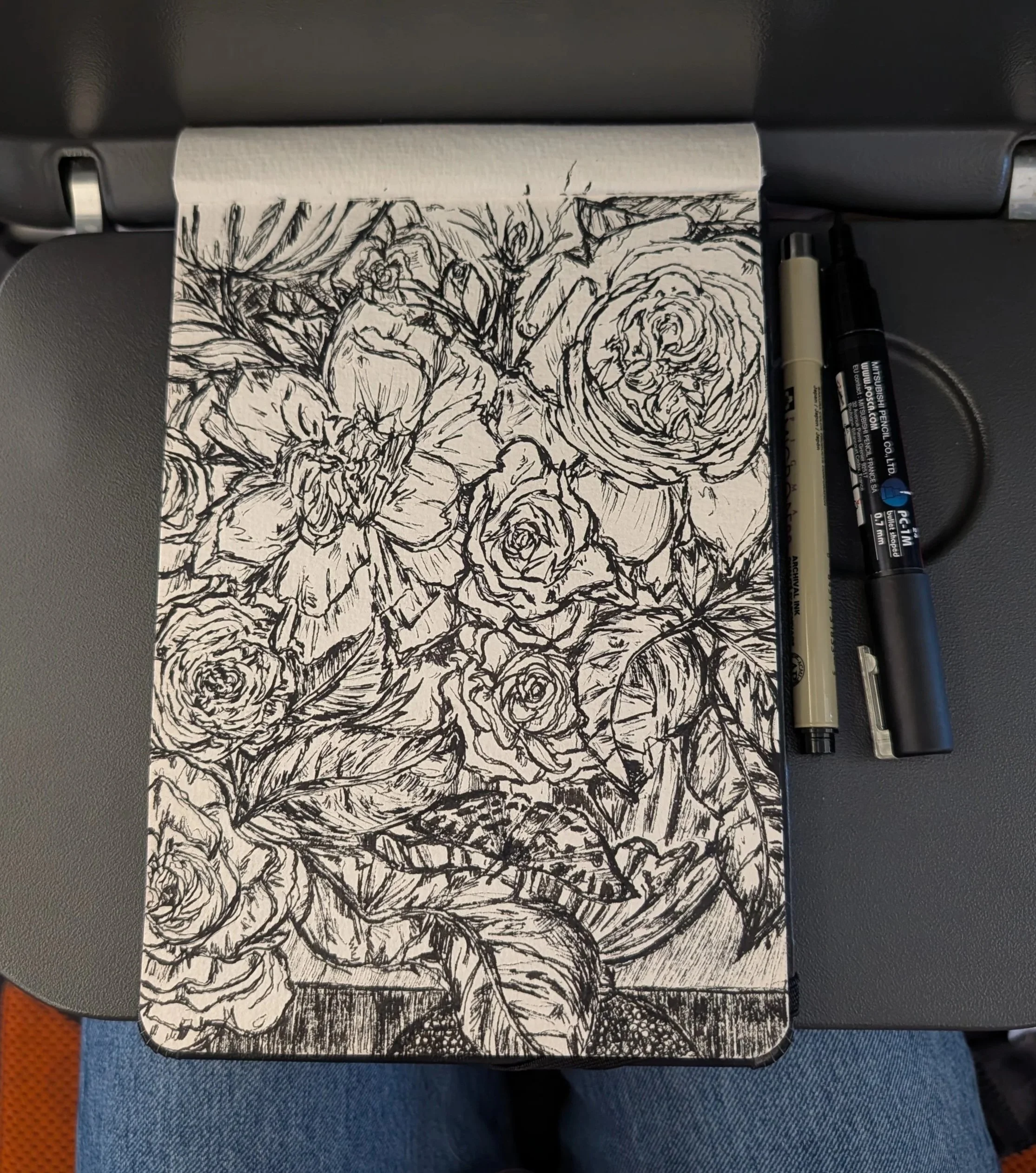 A black and white floral sketch on a small rectangular piece of paper, resting on a train tray table. There are two black markers and one gray marker placed beside it.