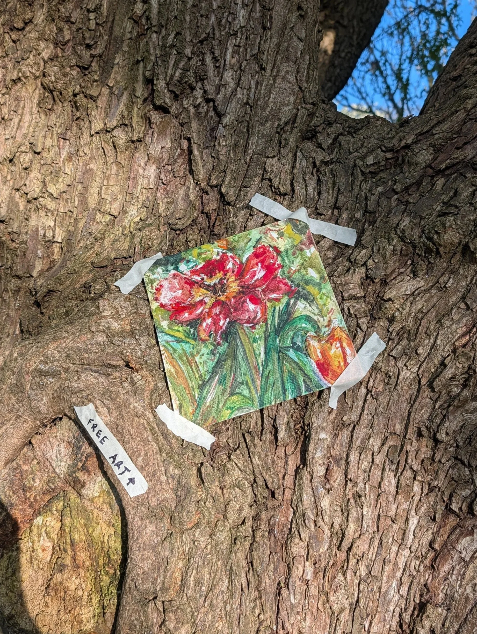 A colorful painted flower on a piece of paper is taped to a tree trunk with a white tape. A small piece of paper with the words 'FREE ART' is attached below.