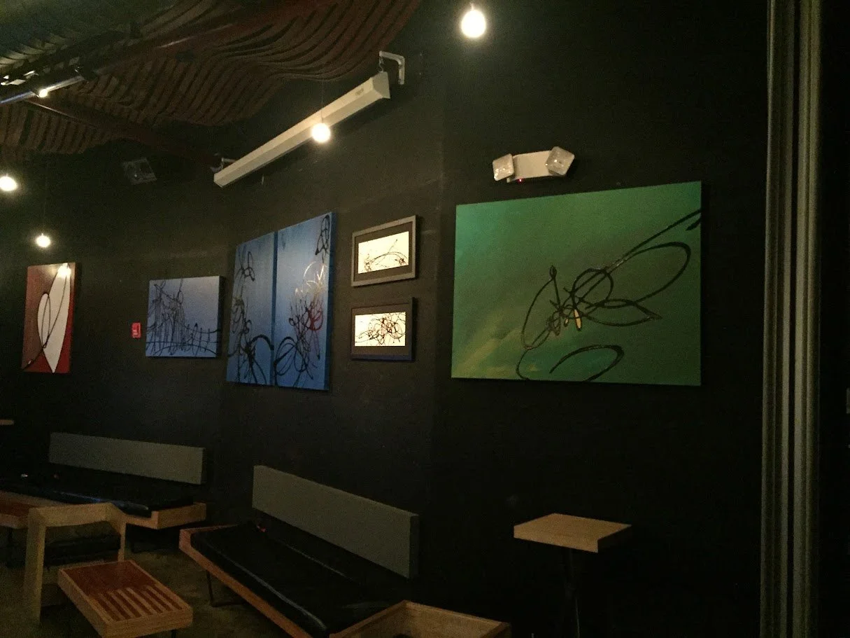 Interior of a dimly lit art gallery with abstract paintings on a black wall, several benches, and small tables.