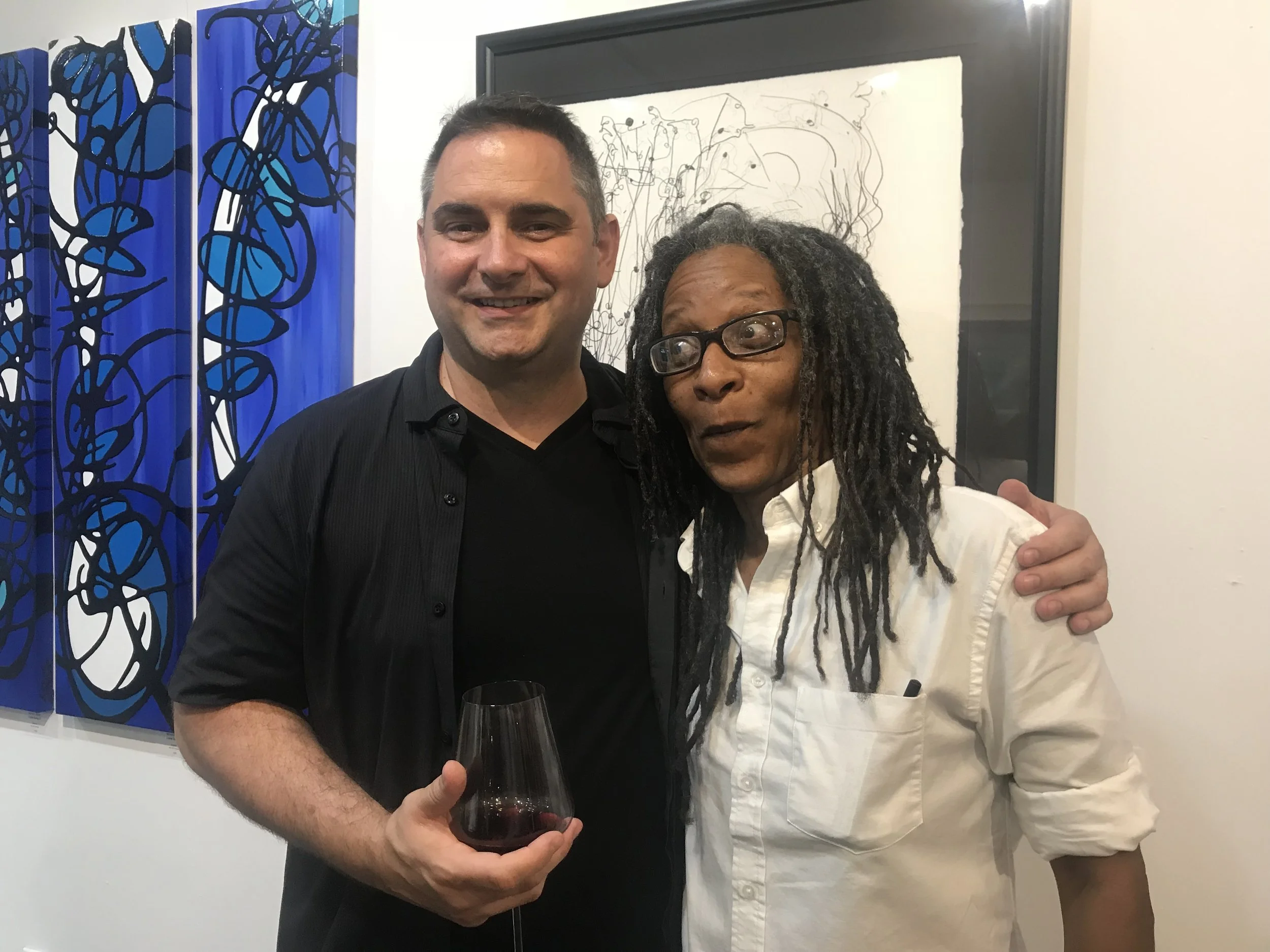 Two people, a man and a woman, standing close together at an art gallery. The man is holding a glass of red wine and is smiling at the camera. The woman is looking to her right with a surprised expression, wearing glasses and a light-colored shirt. Behind them, there are abstract paintings with blue, black, and white colors on display.