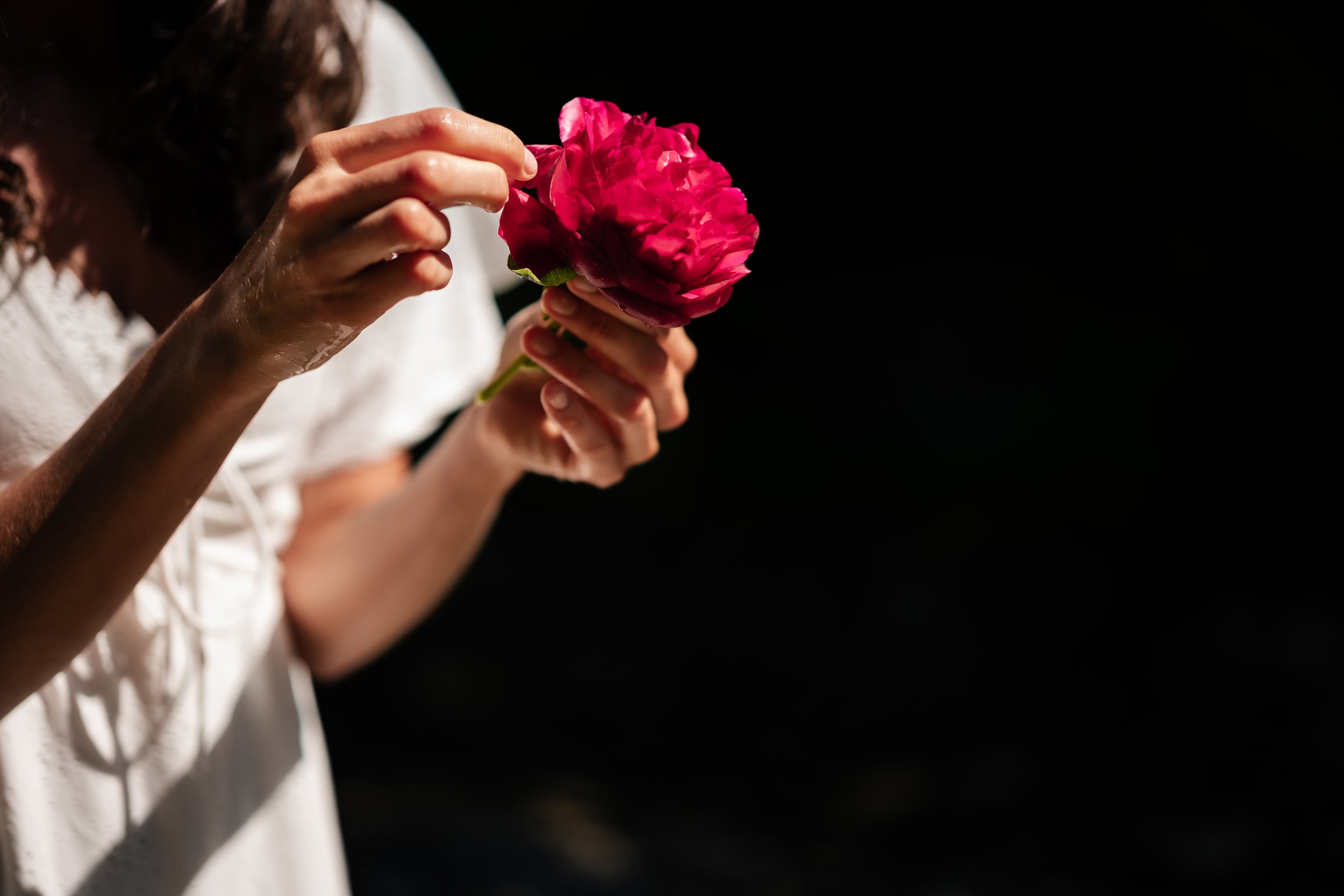 Nahaufnahme einer Person, die eine pinke Blume hält und an ihr schnuppert, vor dunklem Hintergrund.