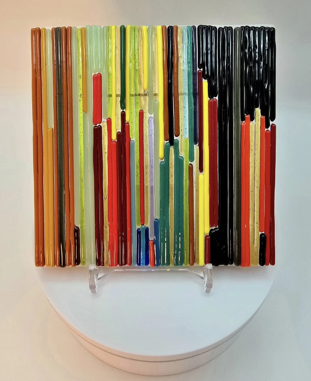 A multicoloured piece of fused glass create from a hundred thin strips of glass and fused together to create one piece of glass still with contours that can be felt by touch. The colours include red, blue, green, yellow, indigo and black.