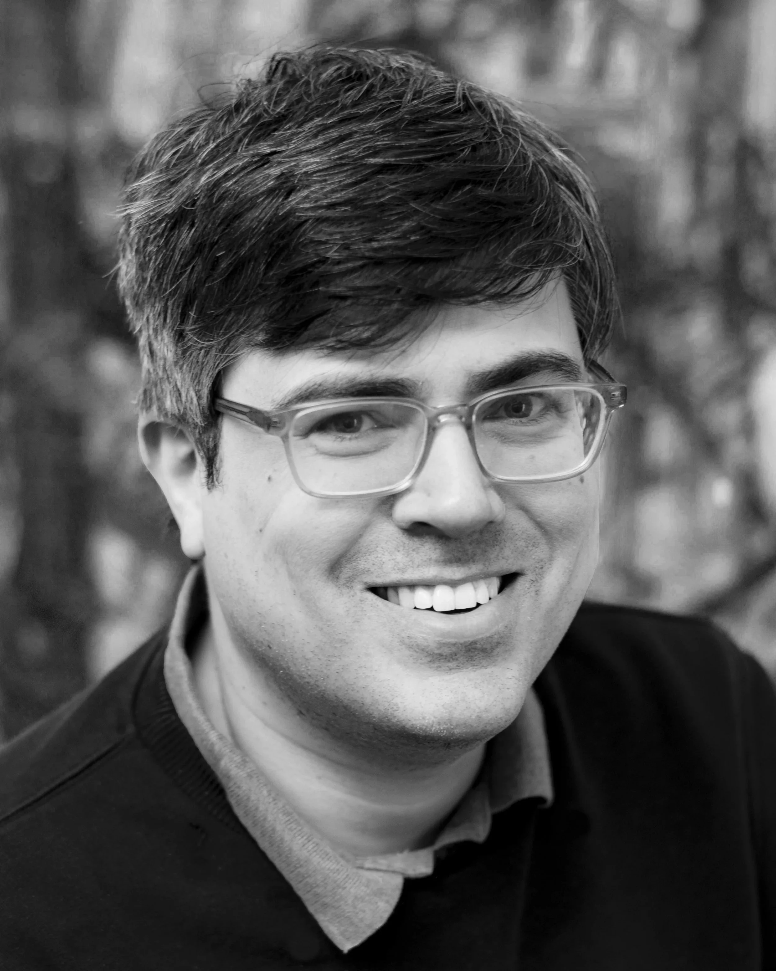 A black-and-white close-up photo of a person's face, showcasing their forehead, eyes with glasses, and a big smile with visible teeth, with short dark hair and wearing a collared shirt and sweater.