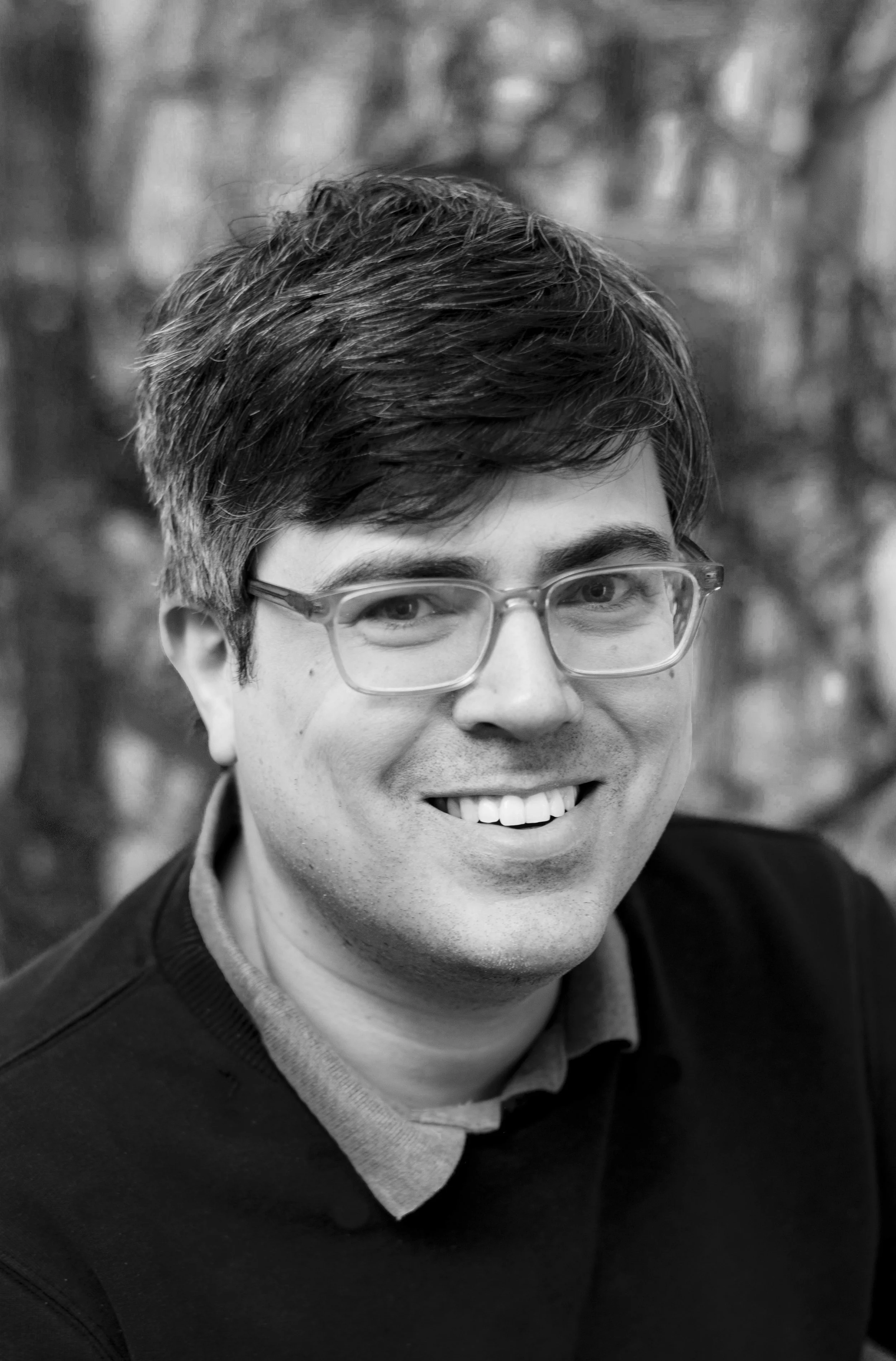 Black and white photo of a smiling man with glasses, short dark hair, dressed in a sweater over a collared shirt, outdoors with blurred background.
