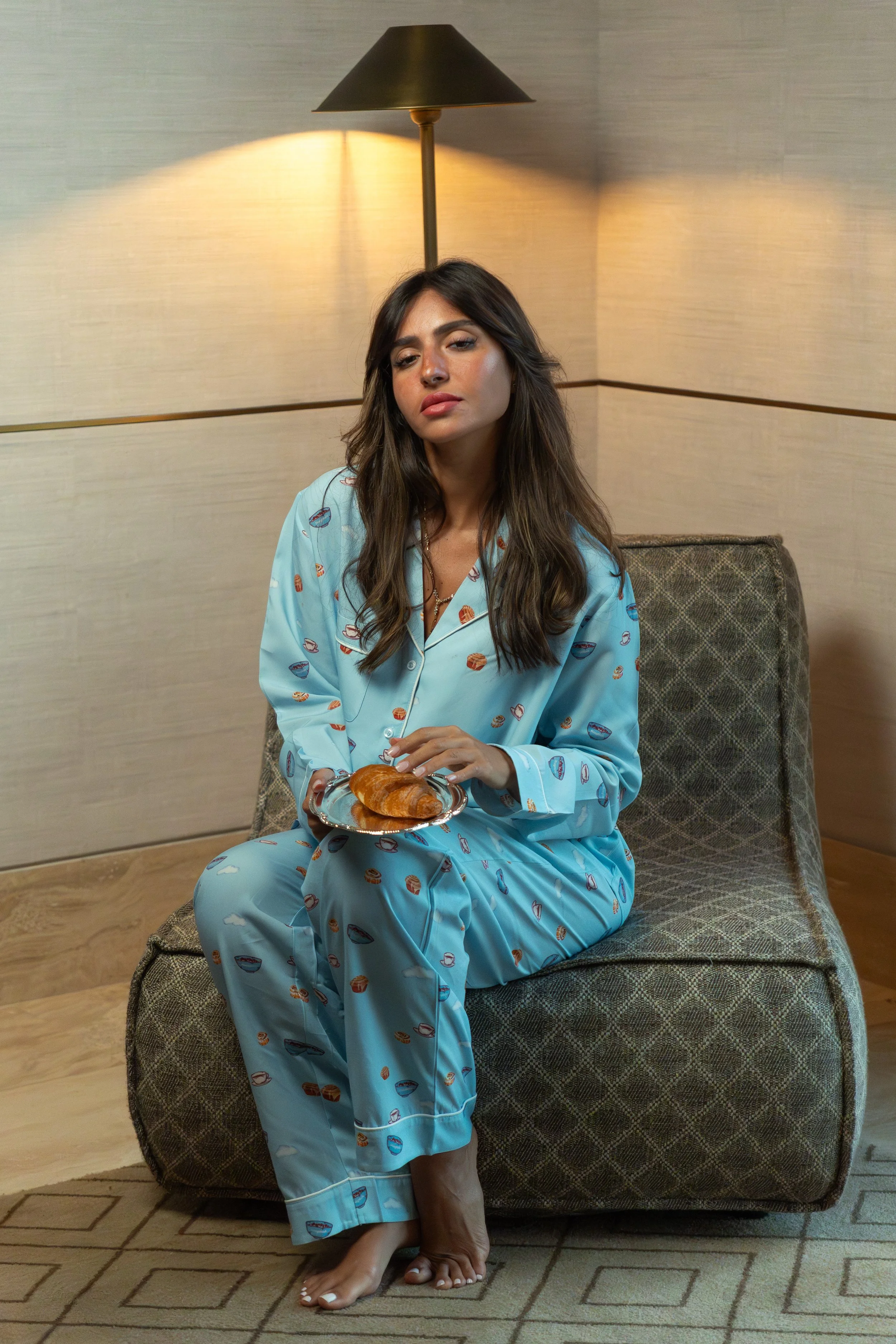 Woman in blue pajamas with a food pattern, sitting cross-legged on a chair, holding a croissant on a silver tray, in a cozy room with beige walls and wooden floor, lit by a tall floor lamp.