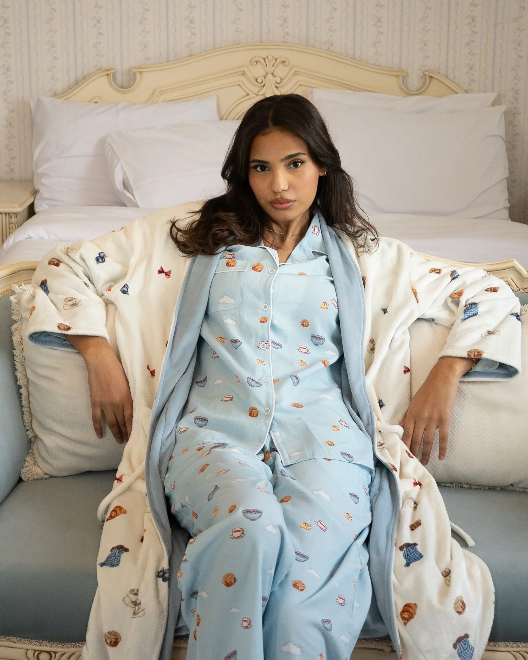 A woman sitting on a bed with a cream-colored headboard and white pillows, wearing pajamas with a food-themed pattern, a matching robe, and looking directly at the camera.