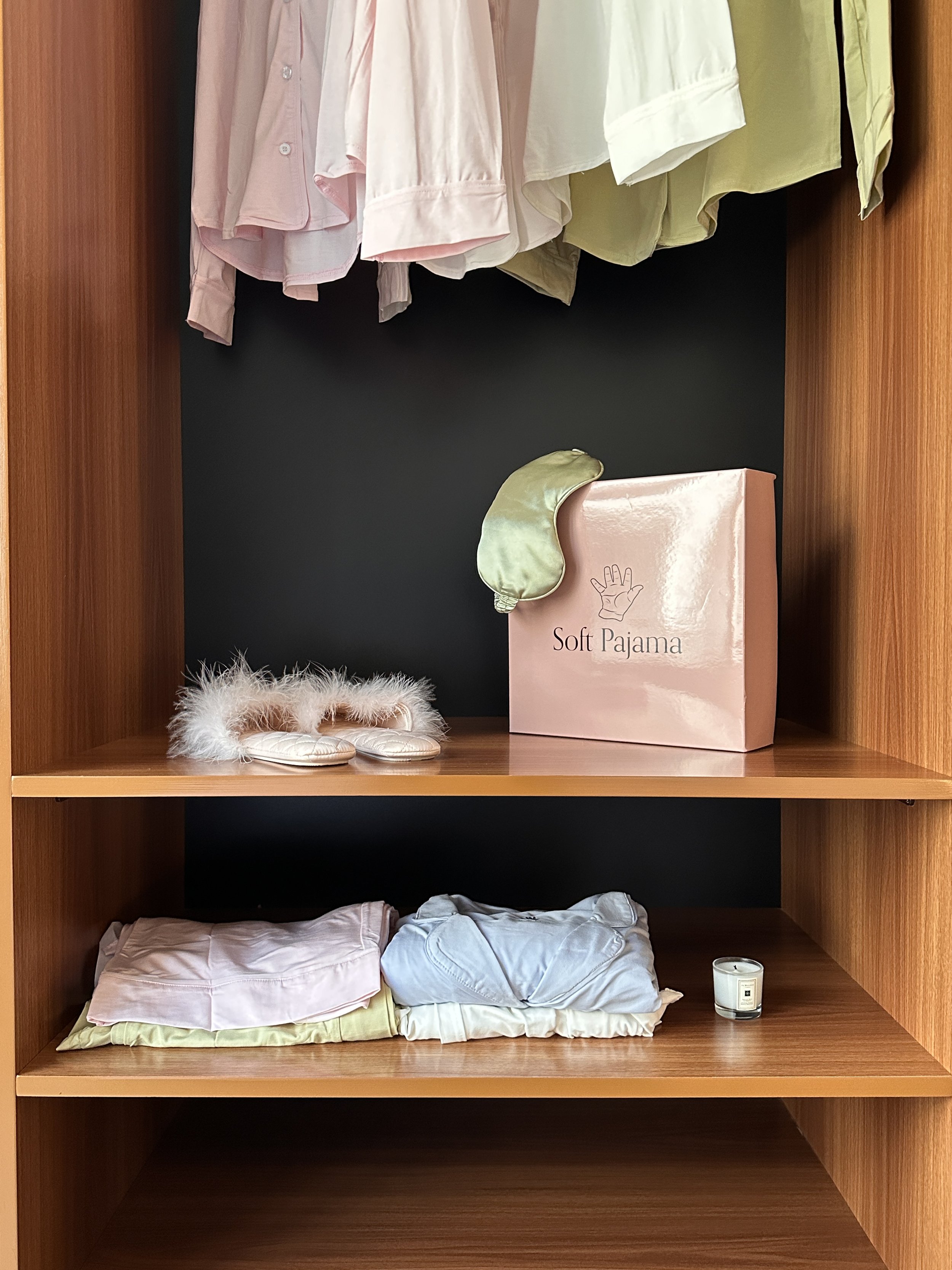 An open wooden wardrobe with shelves, hanging pastel-colored pajamas and shirts, a pink box labeled "Soft Pajama" with a sleep mask on top, folded bedsheets, and a candle.