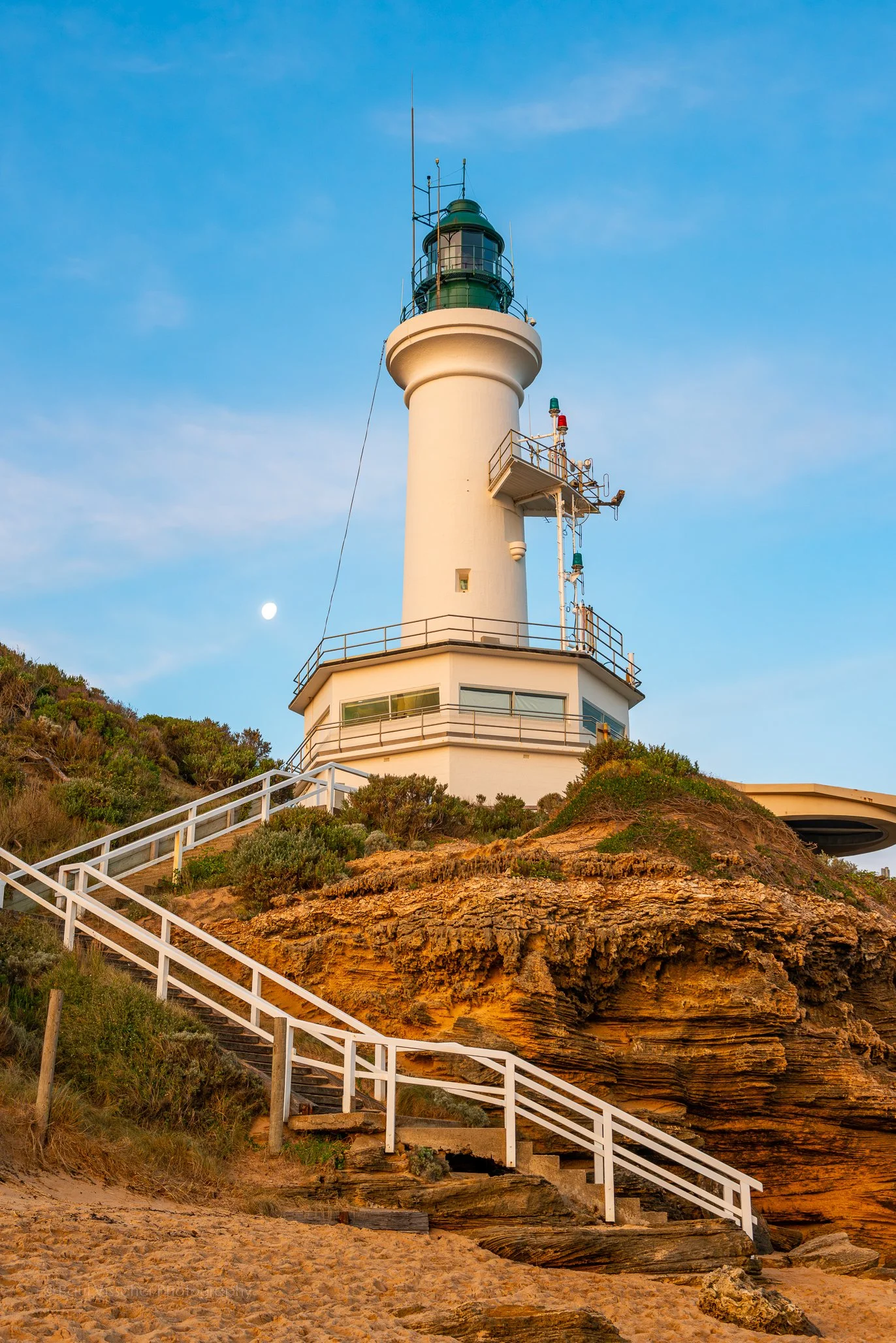 A Point Lonsdale lighthouse on a rocky cliff with stairs leading up to it, under a blue sky with some clouds and a visible moon. Point Lonsdale is on the Bellarine Peninsula in Victoria, Australia. This is an image by photographer, Paul Visscher.