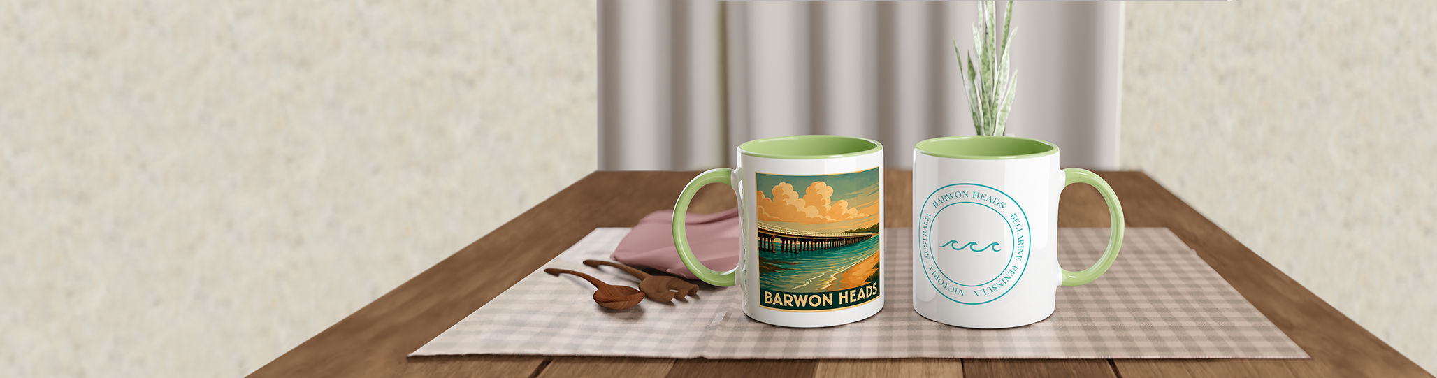 Two coffee mugs with 'Barwon Heads' branding on a wooden table, one with a beach scene and the other with a wave logo, alongside pink and purple napkins, wooden spoons, and a small plant in the background.