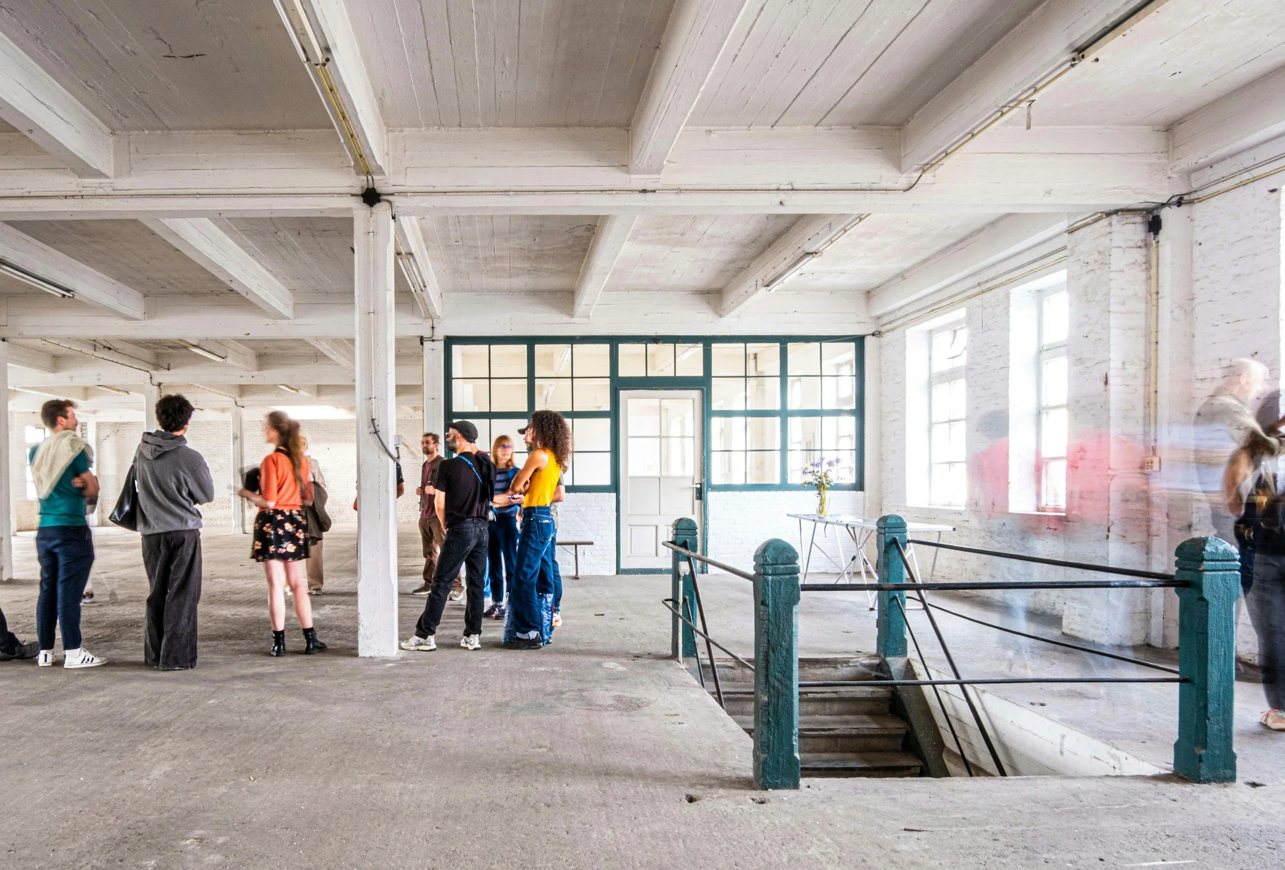 People gathering inside an industrial-style space with high ceilings, white brick walls, large windows, and concrete flooring.