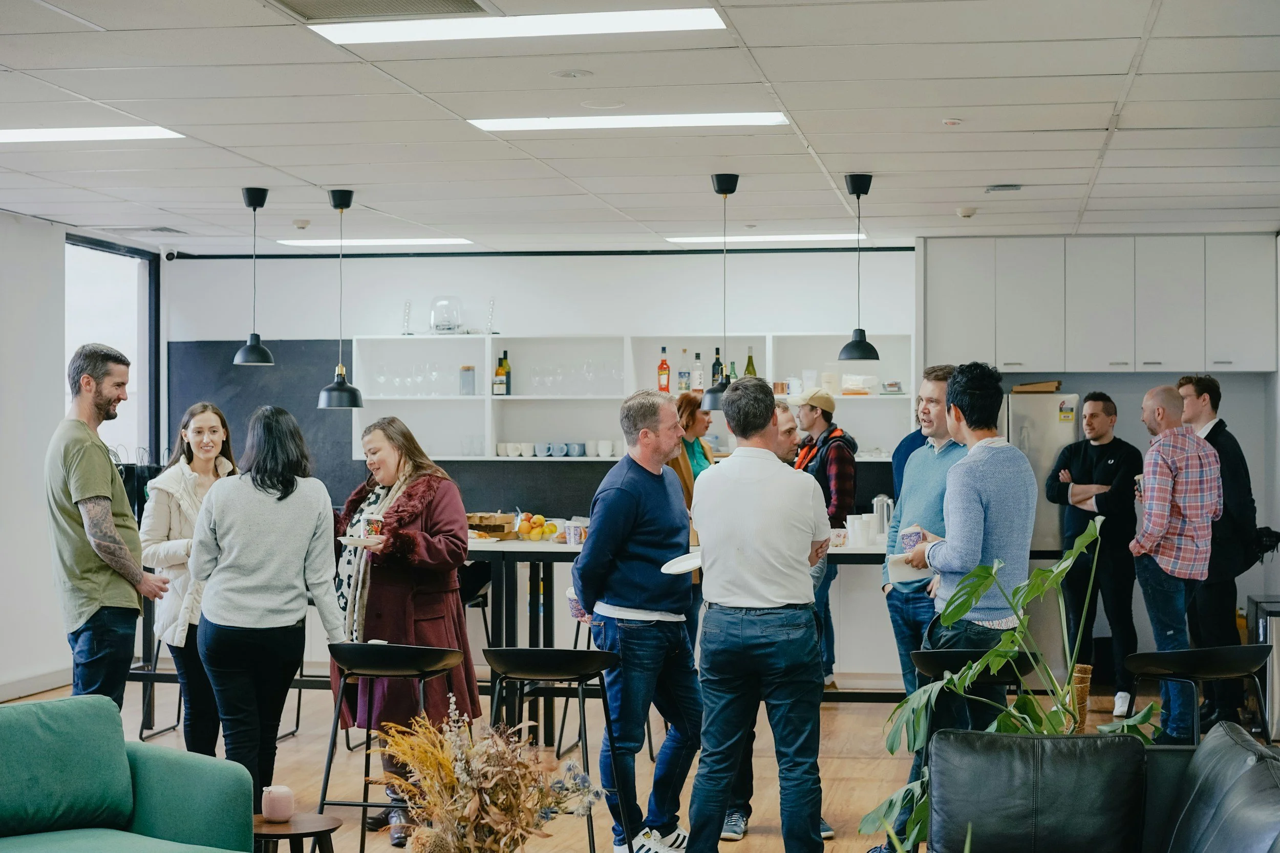 Group of people socializing in a modern kitchen or lounge area, some standing and some engaged in conversation, with a kitchen counter and shelves with bottles and glasses in the background.