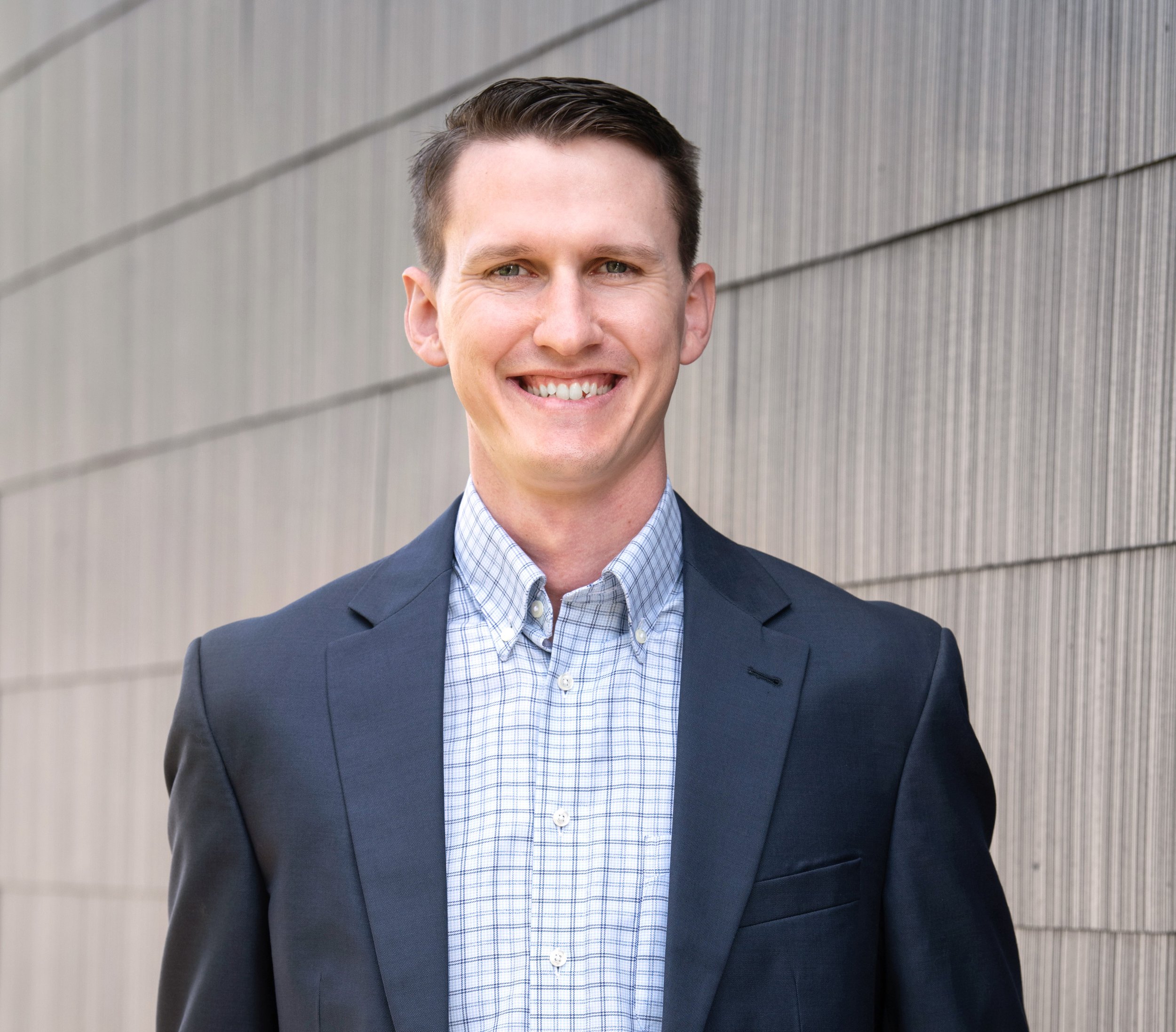 A headshot of a smiling man with short brown hair wearing a dark suit and blue checkered shirt, standing outdoors against a modern gray metal wall.