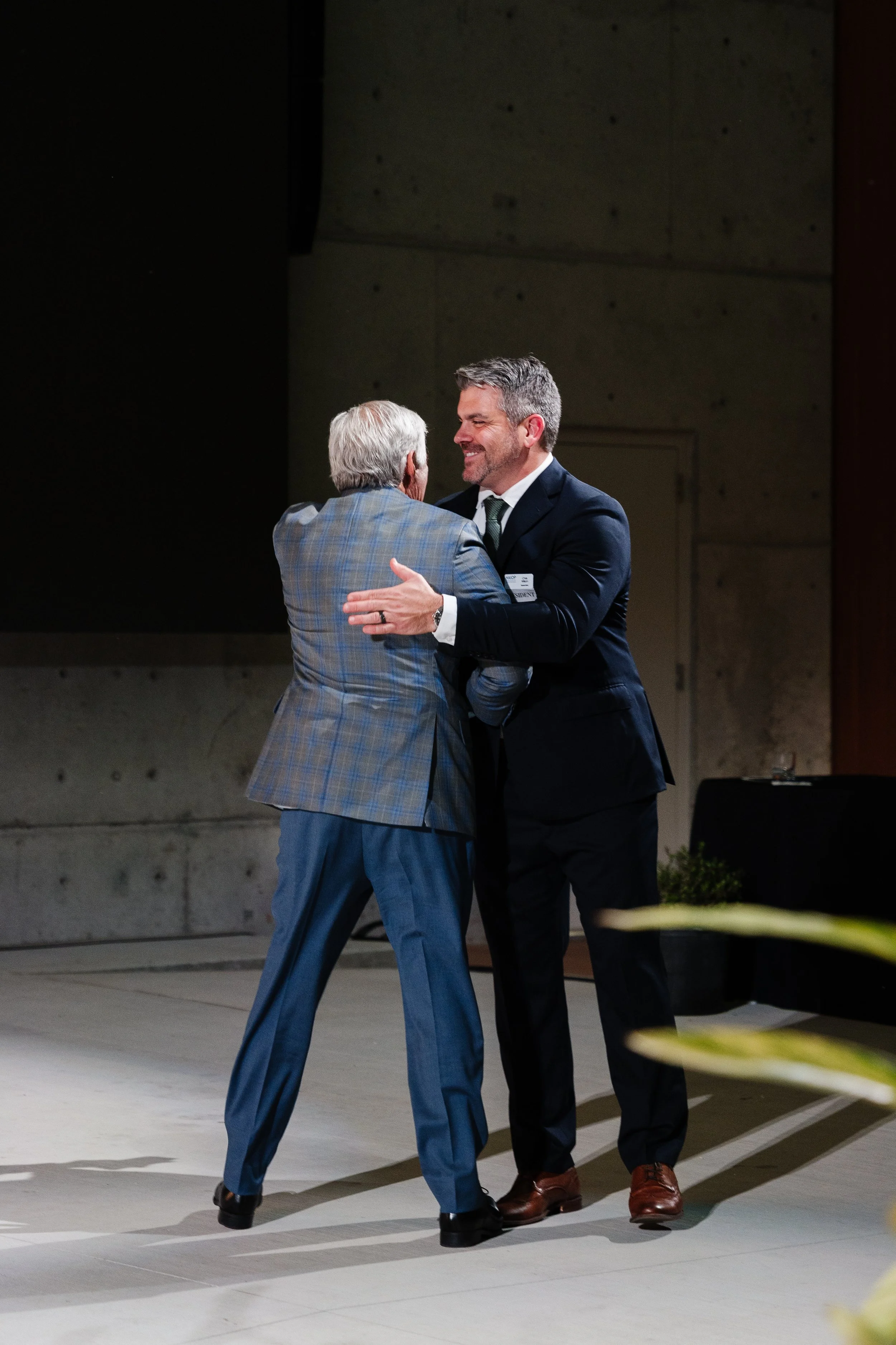 Two men in suits greeting each other warmly and smiling in a hallway or conference space.