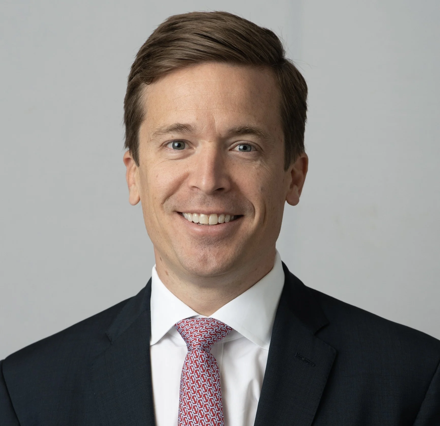 Close-up of a smiling man wearing a dark suit, white shirt, and red patterned tie, against a plain gray background.