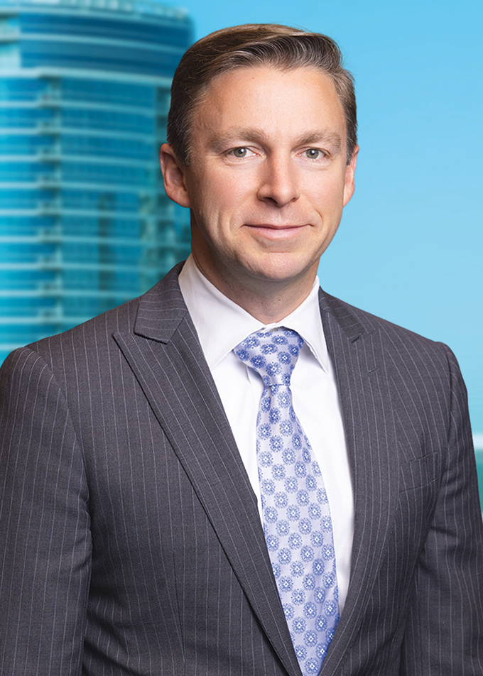 A professional man in a dark pinstripe suit with a light blue patterned tie, standing in front of a blue background with blurred skyscrapers.