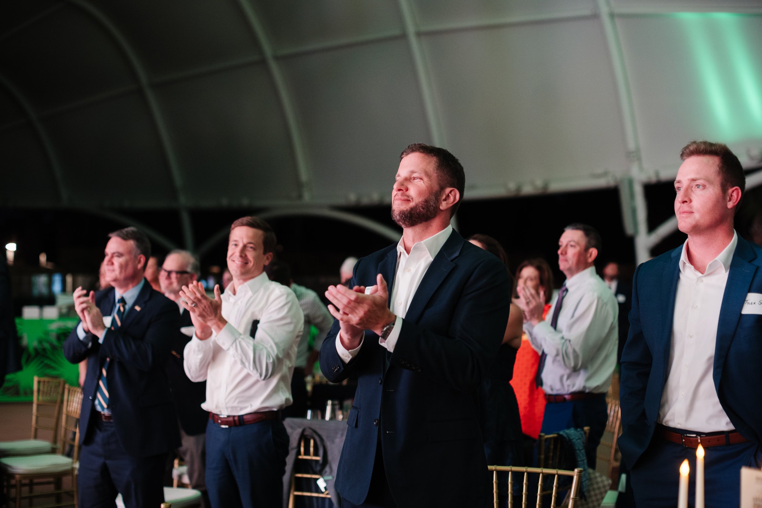 A group of people at an indoor event, standing and clapping, with some smiling and others with serious expressions, dressed in formal attire.