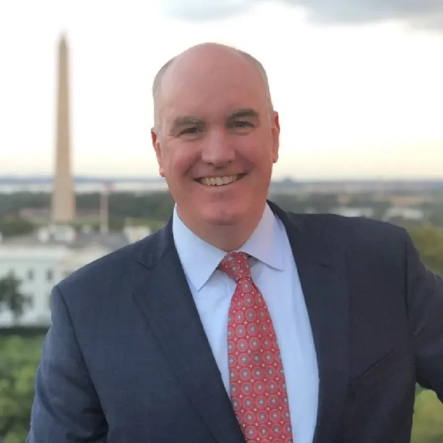 A smiling man in a suit and red-patterned tie standing outdoors with a cityscape and the Washington Monument in the background.