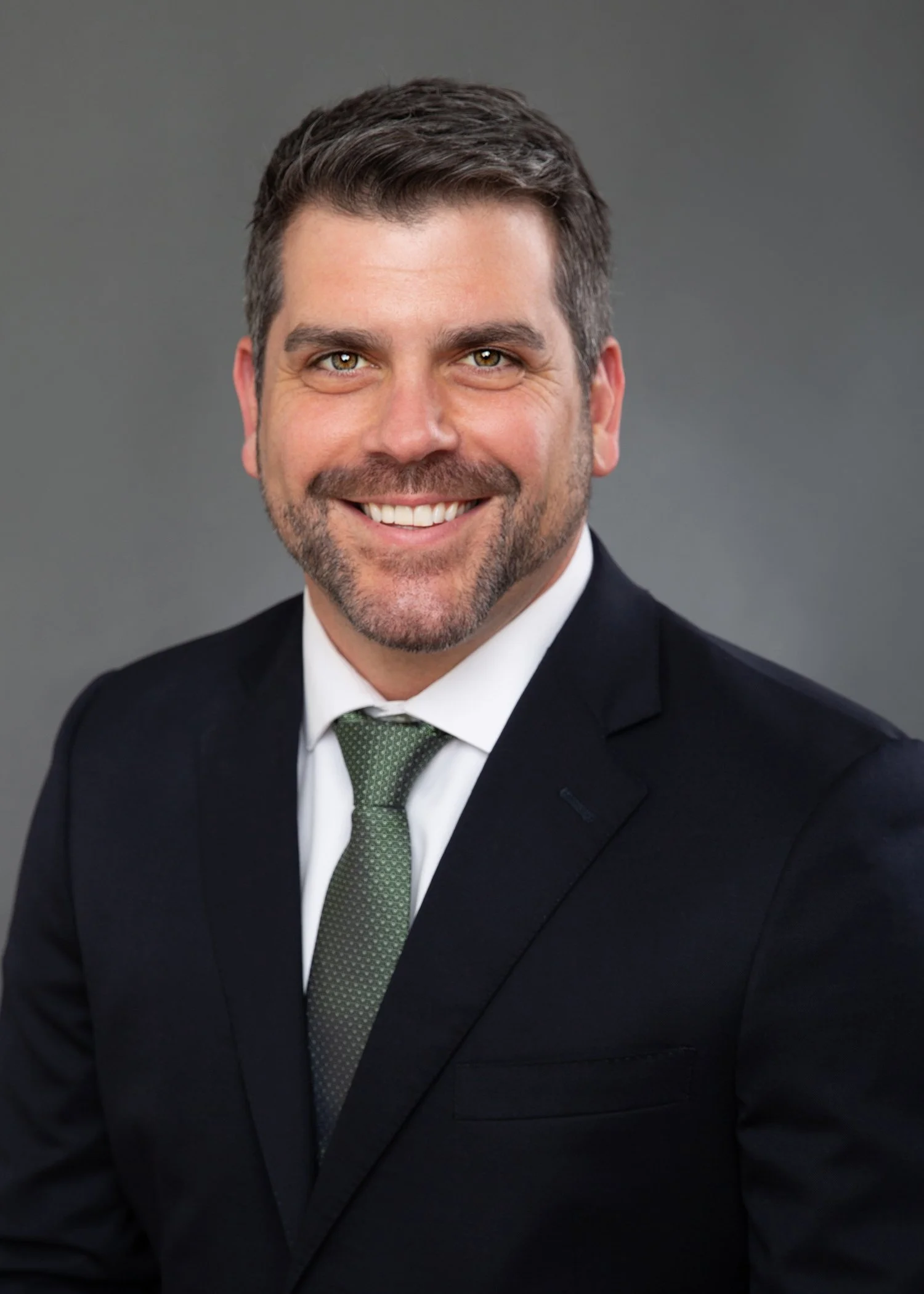 Portrait of a smiling man in a suit, wearing a white shirt and a green tie, against a plain gray background.