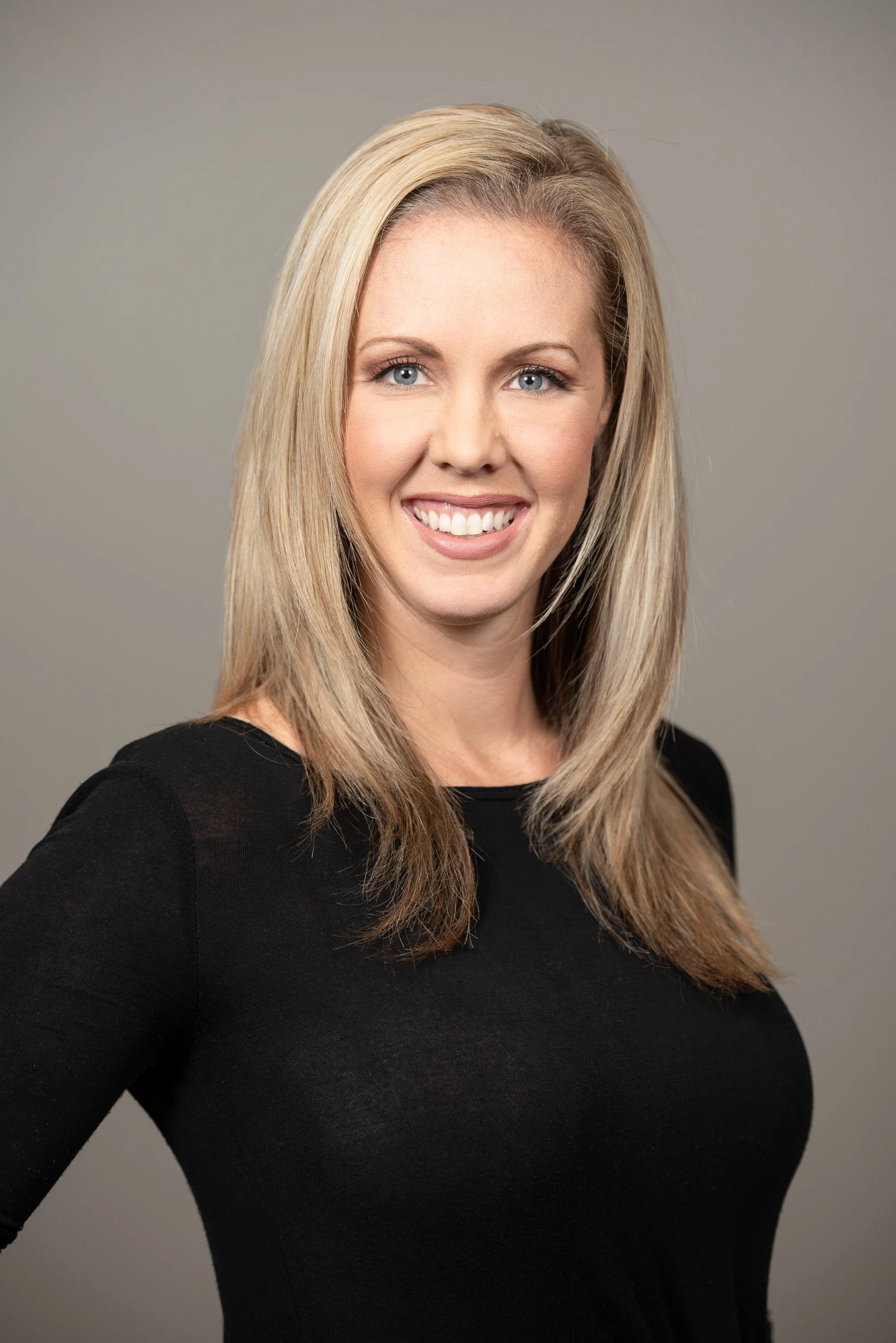 Portrait of a smiling blonde woman with blue eyes wearing a black top against a gray background.