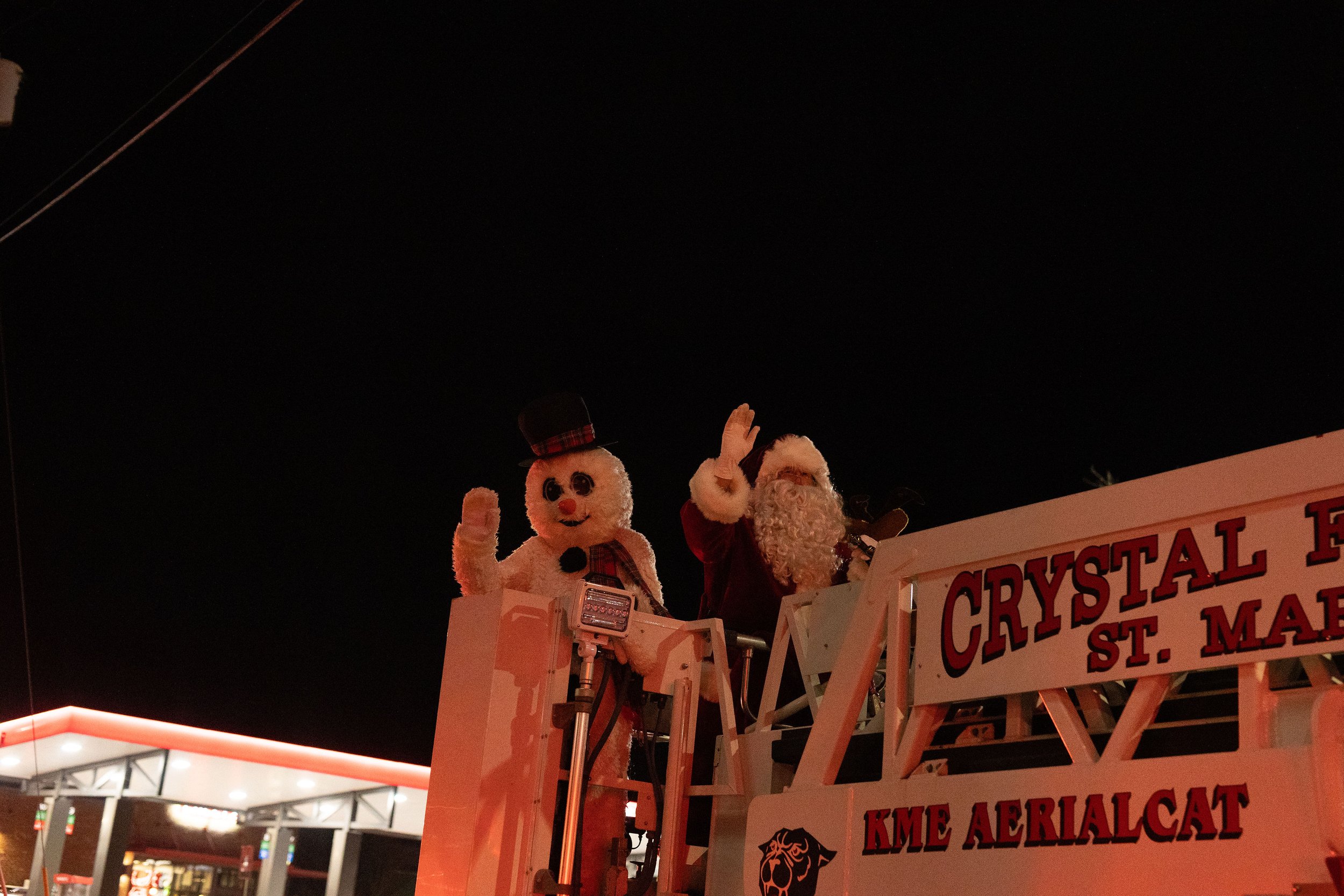 Santa Claus and a snowman figure on a float during a nighttime Christmas parade.