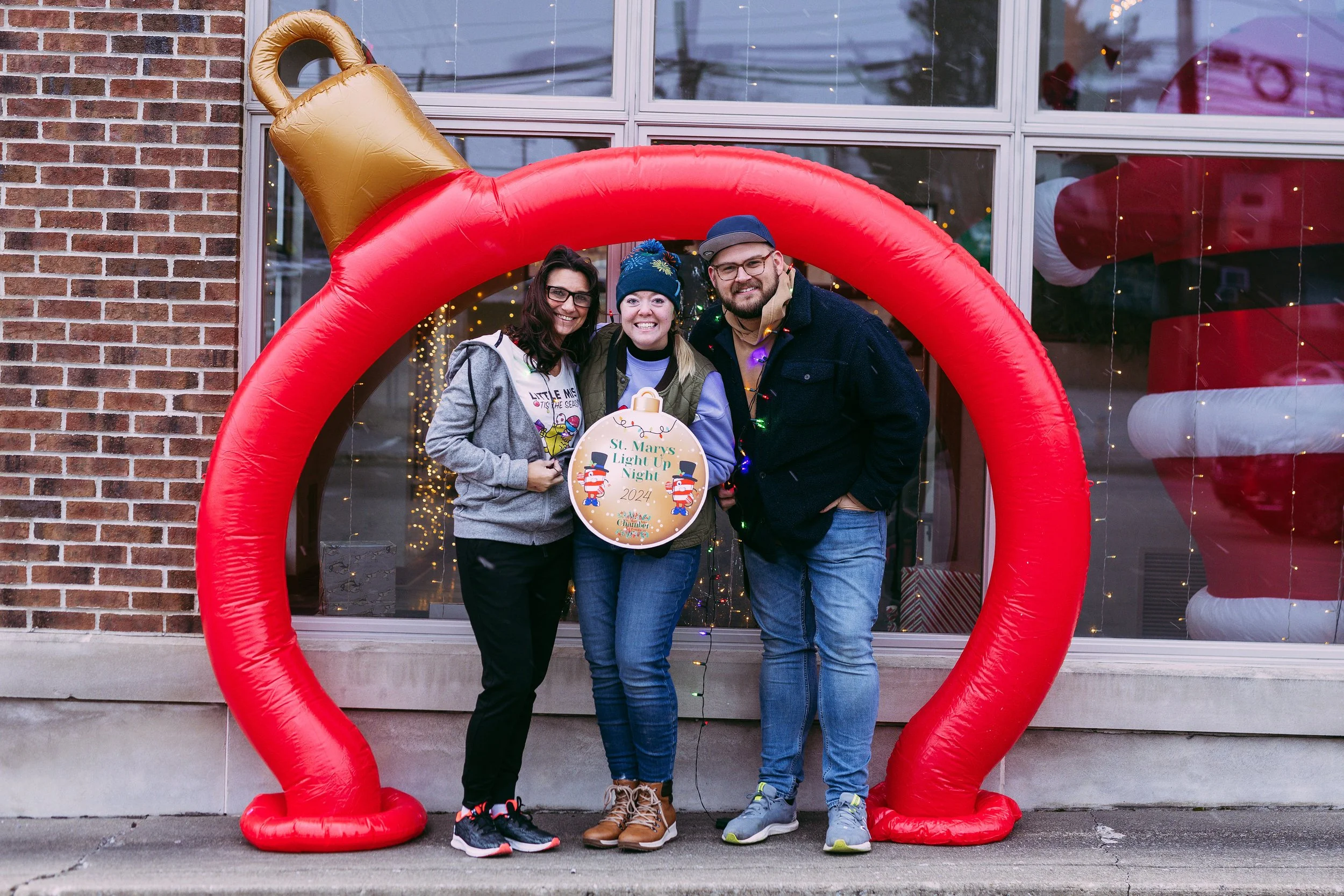 Three people smiling and posing for a photo inside an inflatable red heart-shaped decoration during a holiday celebration in front of a building with a glass window decorated with string lights. They are holding a sign that reads 'St. Mary's Light Up