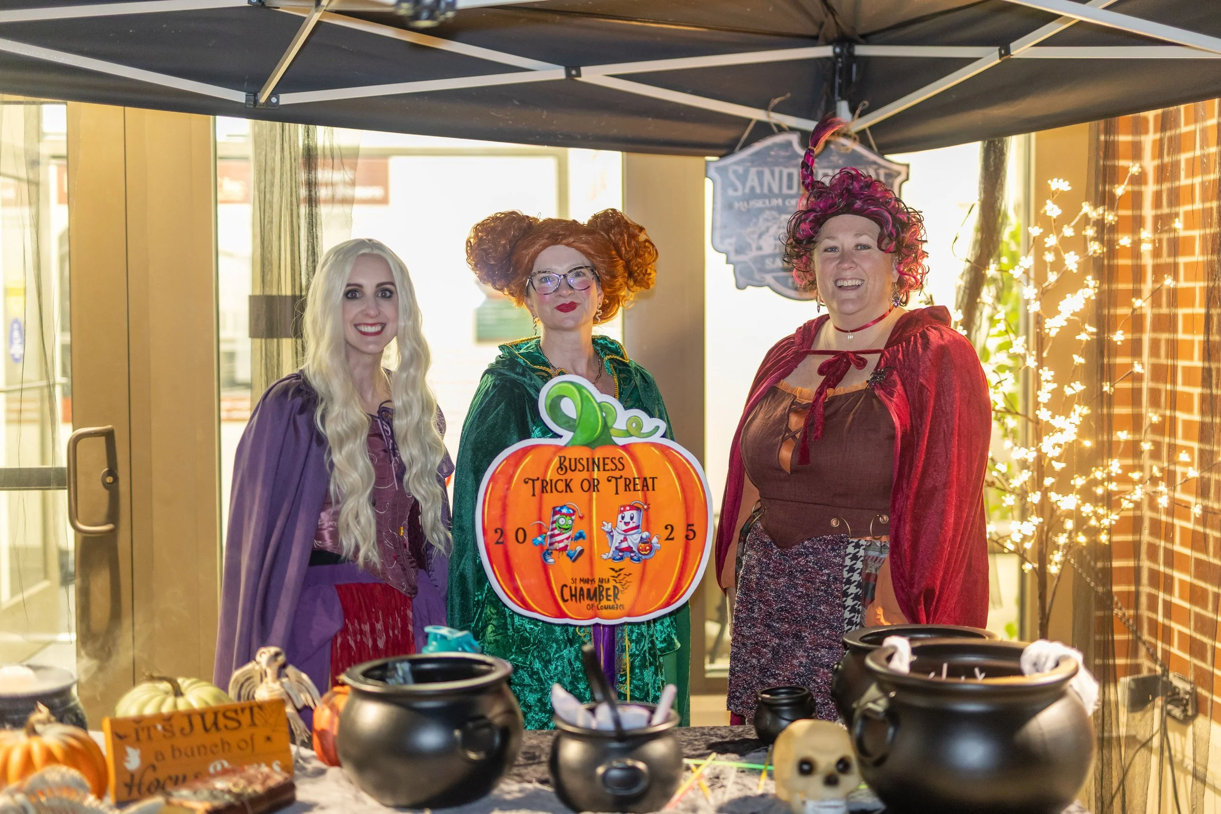 Three women dressed as witches standing behind a table with Halloween decorations at an indoor event, smiling. One woman has long blonde hair, the second has curly red hair and glasses, and the third has curly dark hair with pink highlights. A sign o