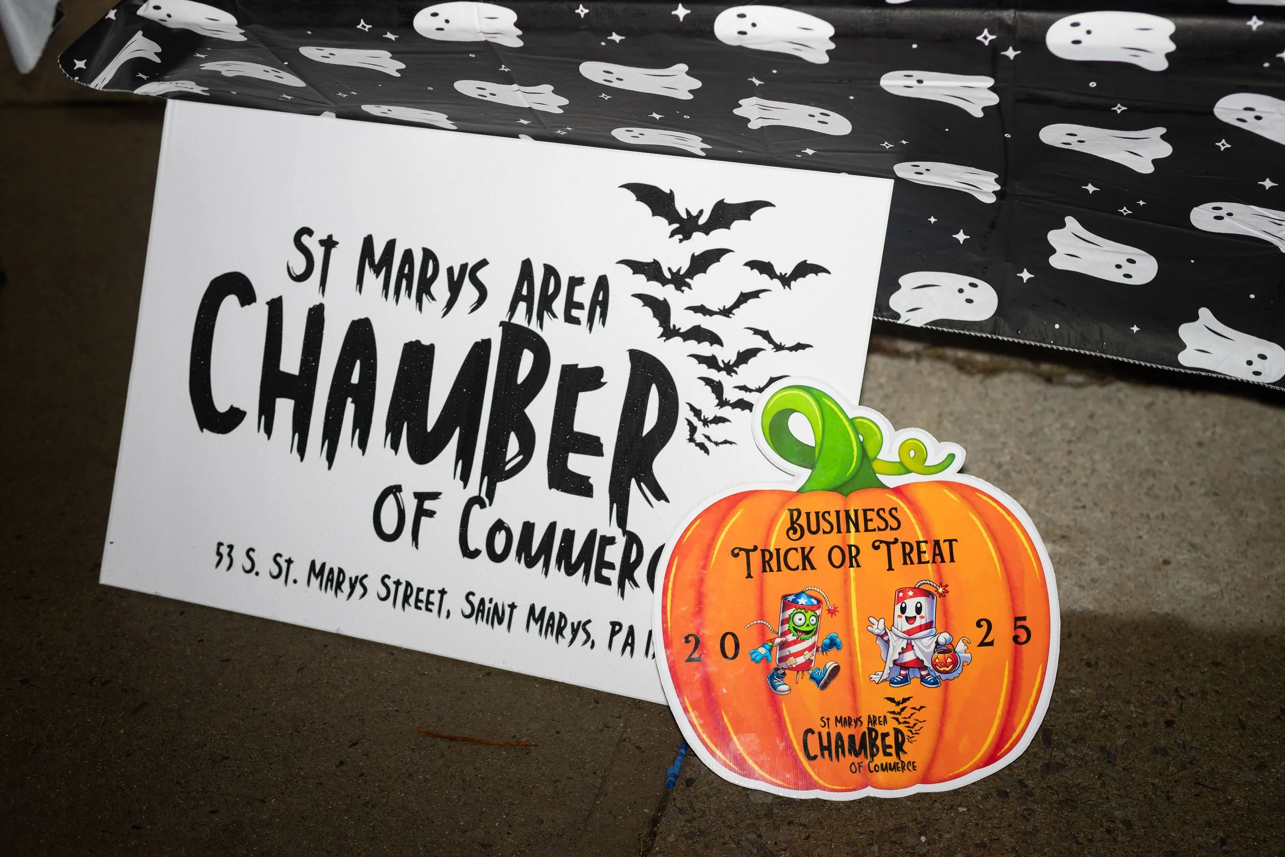 A Halloween-themed event sign on a sidewalk with a black background decorated with white ghost and star patterns, advertising the St. Marys Area Chamber of Commerce. A pumpkin-shaped sign with Halloween decorations and the words "Business Trick or Tr