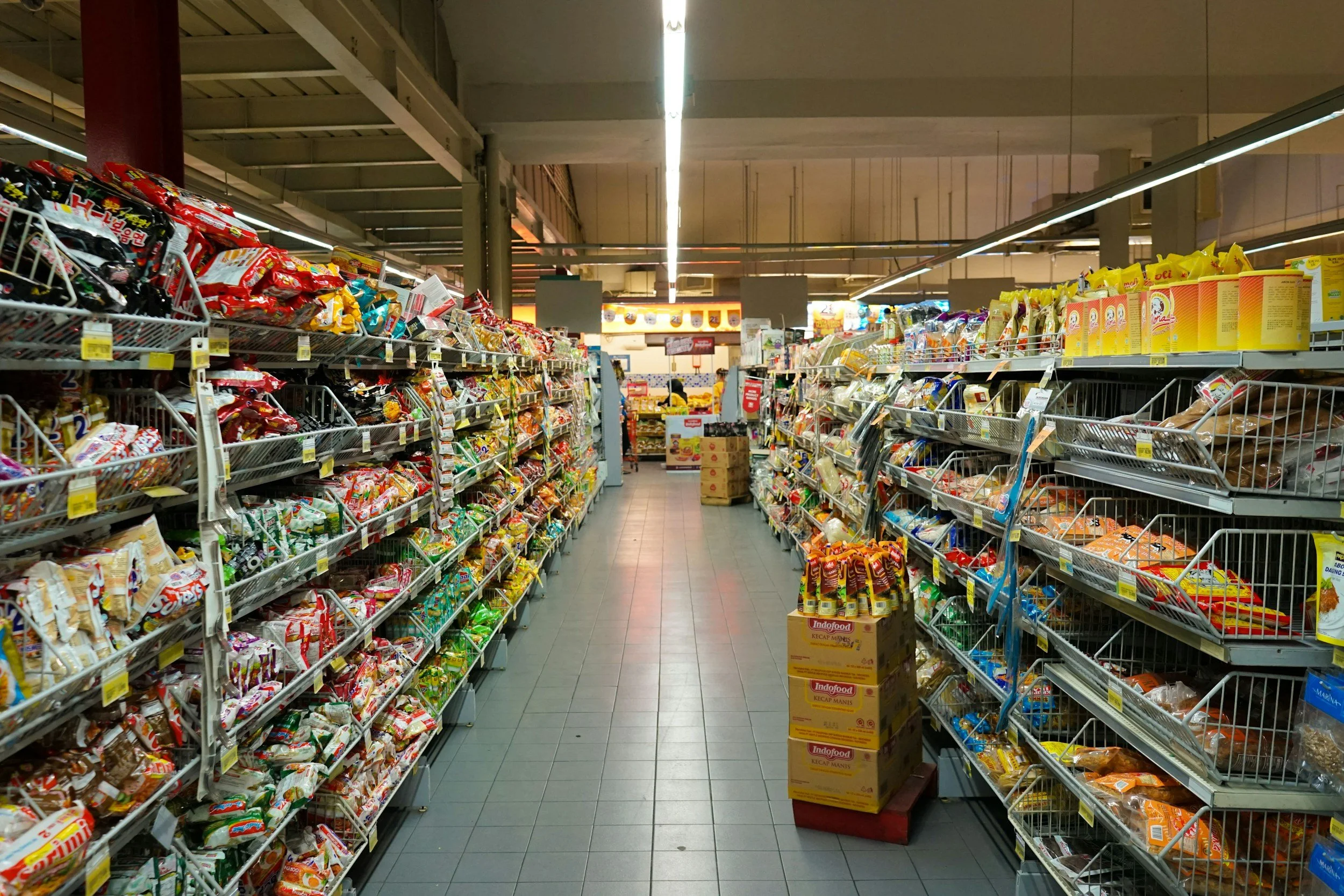 Aisle in a grocery store with shelves stocked with snack foods, including chips and instant noodles, with boxes stacked on the floor in the foreground.