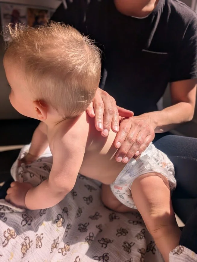 Un bébé allongé sur le ventre, recevant un massage ou une thérapie par une personne adulte, probablement un thérapeute, dans une pièce intérieure.