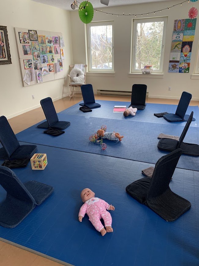 Pièce de yoga ou salle de réunion avec tapis bleu, chaises renversées, jouets pour bébé et décorations colorées suspendues au plafond, fenêtres laissant entrer la lumière.