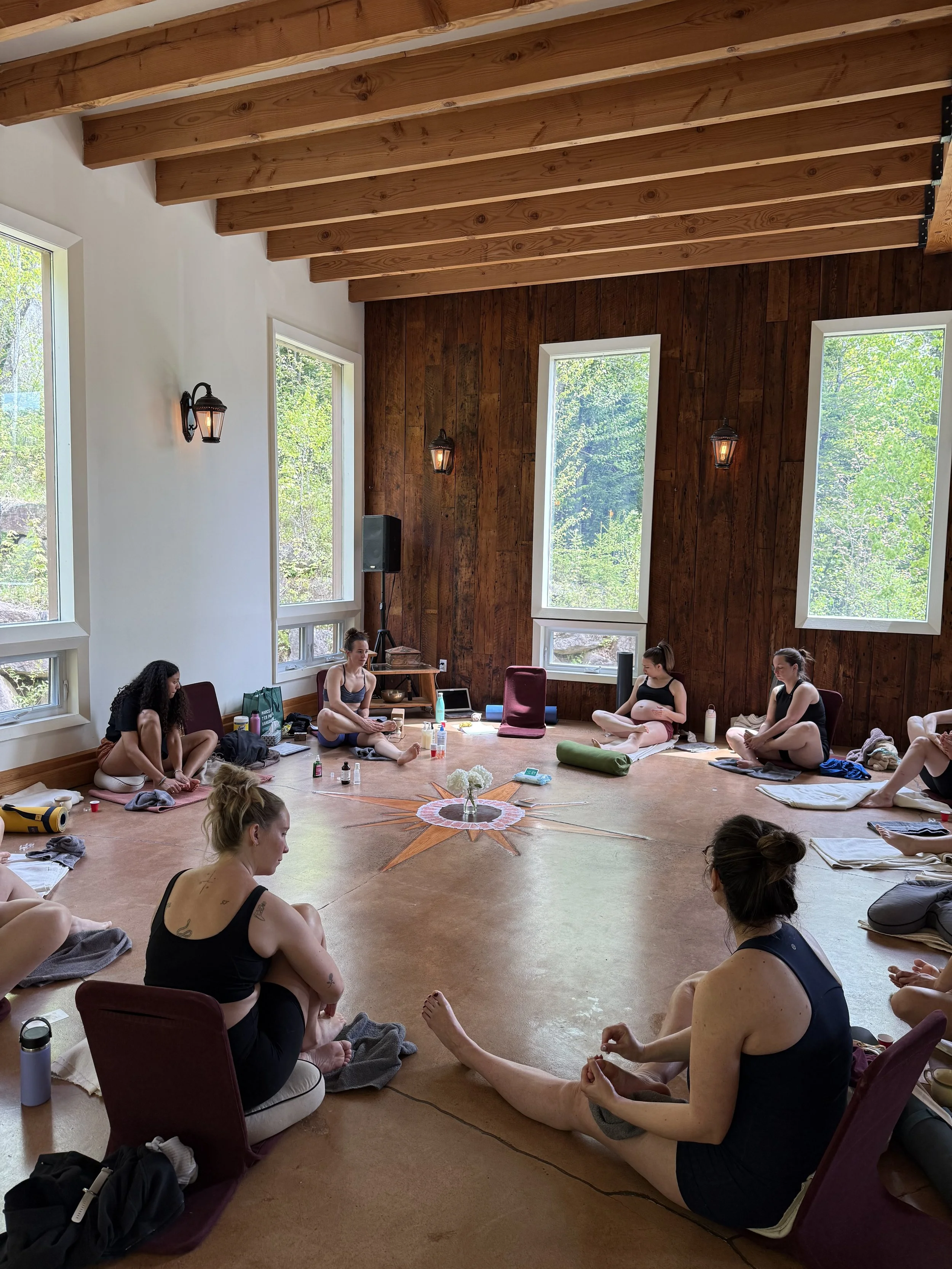 Groupe de personnes pratiquant le yoga ou la méditation dans une salle en bois avec de grandes fenêtres et des lampes suspendues.